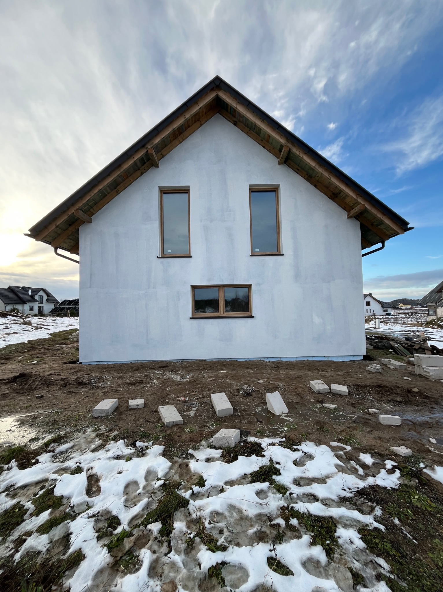 Surowa elewacja nowo wybudowanego domu jednorodzinnego w trakcie prac, z widocznymi bloczkami fundamentowymi i resztkami śniegu na ziemi.