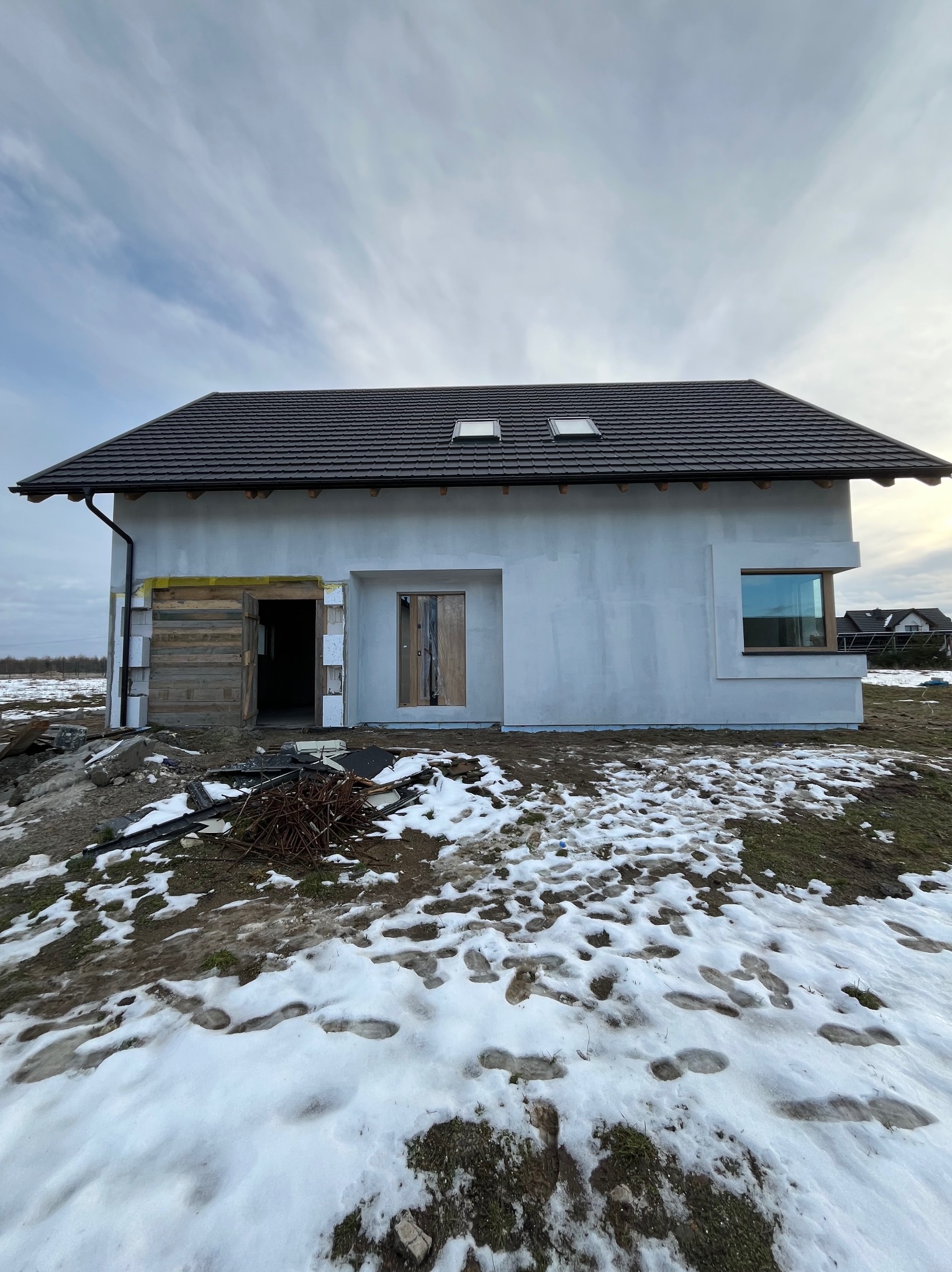 Surowy stan budynku jednorodzinnego z szarą elewacją, widoczne otwory okienne i drzwiowe, częściowo ocieplone, na pierwszym planie resztki śniegu i błoto.