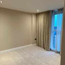 MC Service - Pusty pokój z beżowymi ścianami, jasną wykładziną dywanową i zasłonami w oknie, widoczny fragment budynku za oknem.