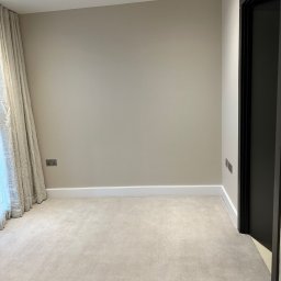 MC Service - Pusty pokój z beżową ścianą, kremowym dywanem, białymi listwami przypodłogowymi i czarnymi drzwiami, oświetlony punktowo.