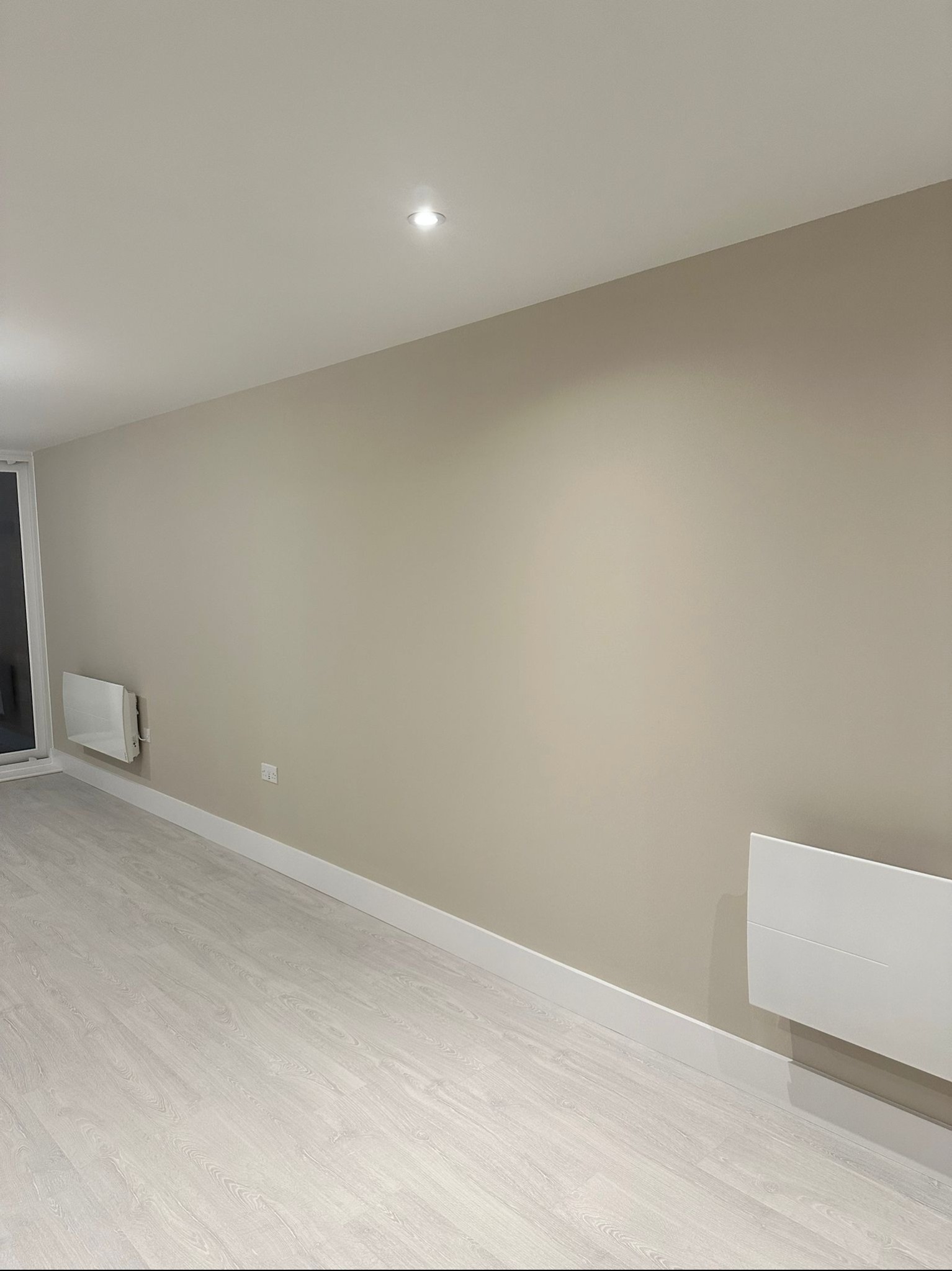 Jasne wnętrze po malowaniu ścian na beżowo, z białymi panelami podłogowymi i grzejnikiem elektrycznym na ścianie.