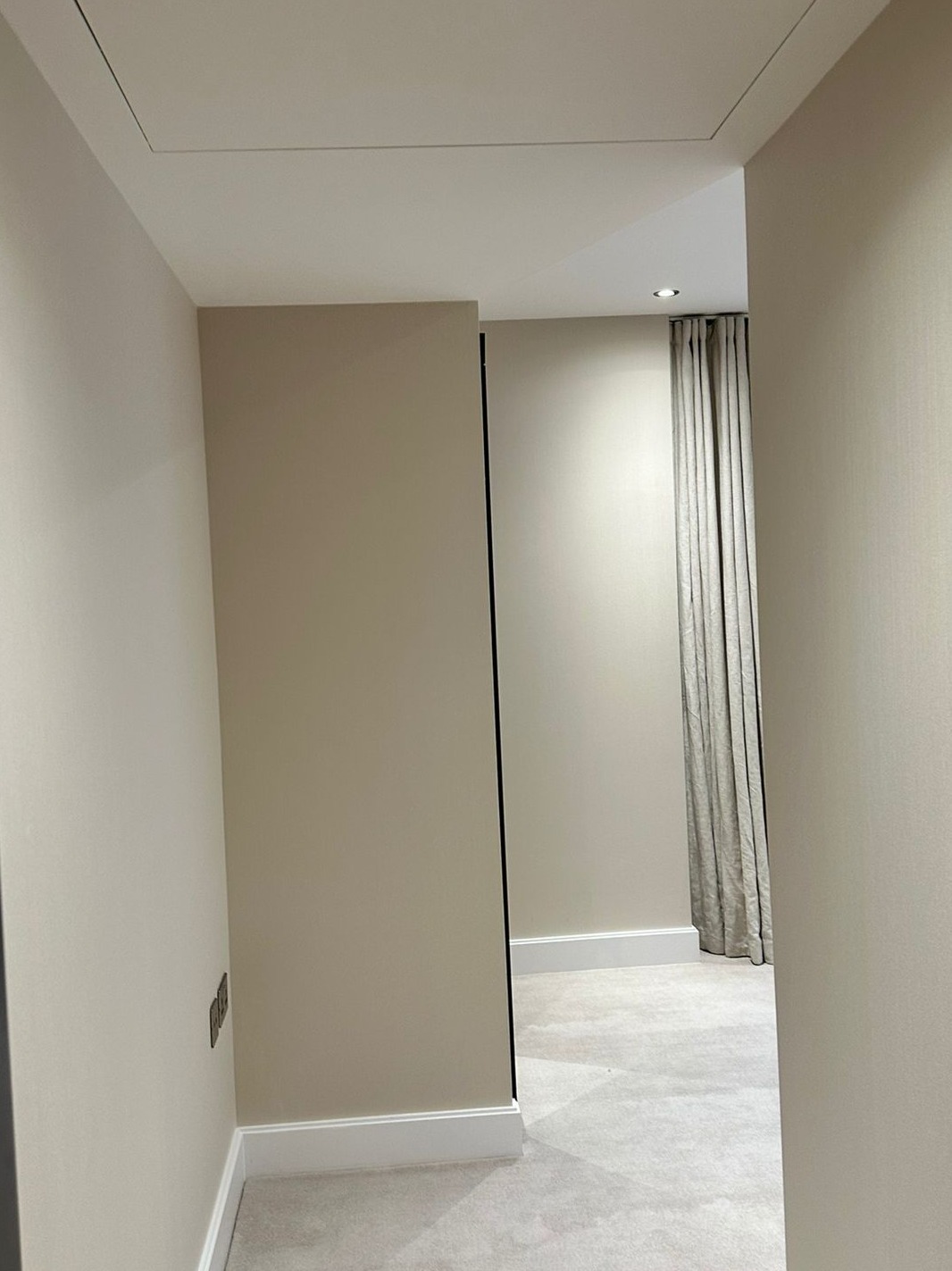 Korytarz z beżowymi ścianami, listwami przypodłogowymi, ukrytym włazem w suficie i widokiem na pokój z zasłonami i wykładziną.
