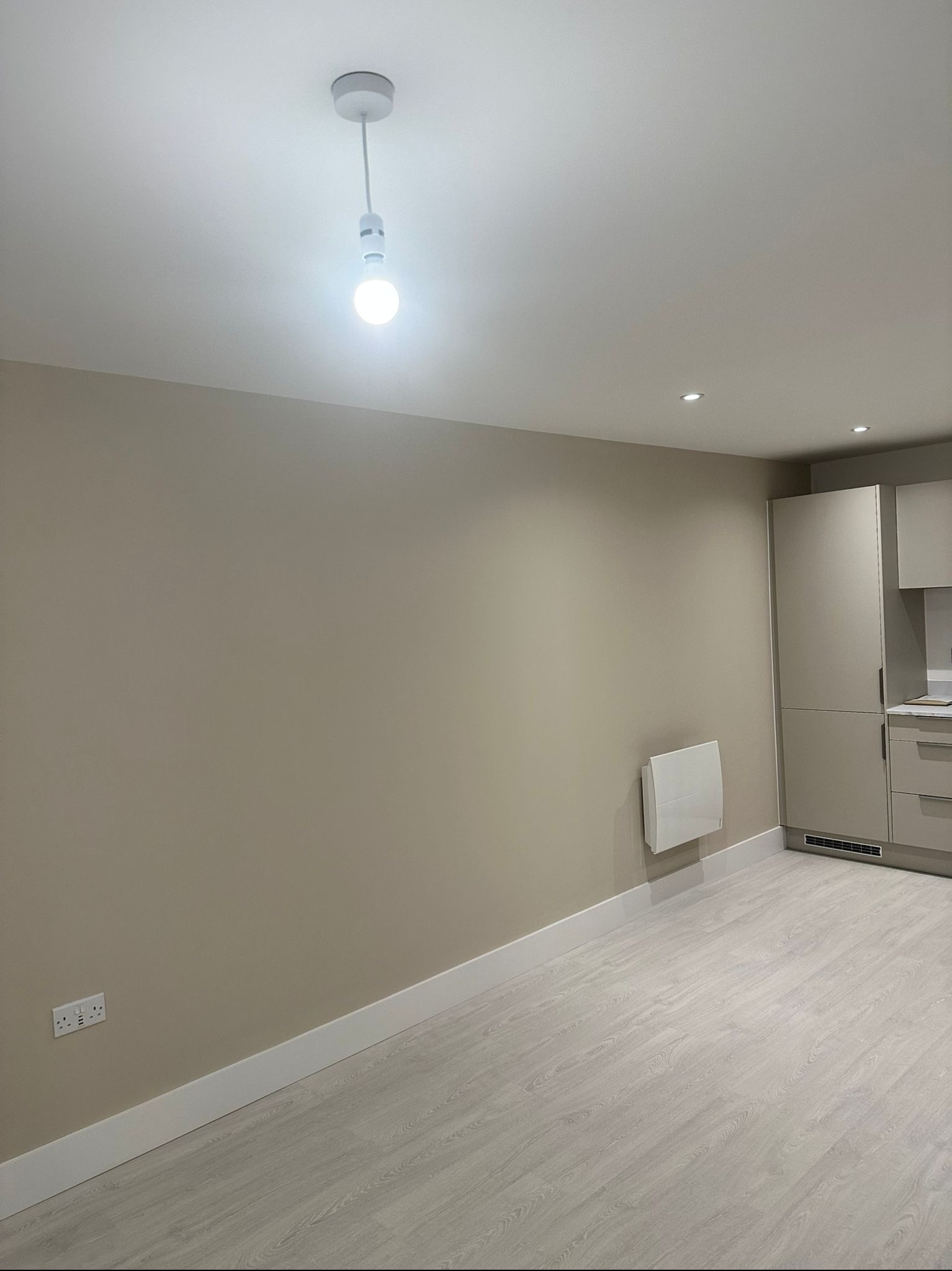 Wnętrze pokoju po remoncie z beżowymi ścianami, białymi listwami przypodłogowymi, panelami podłogowymi w odcieniach szarości oraz fragmentem zabudowy kuchennej w tle.