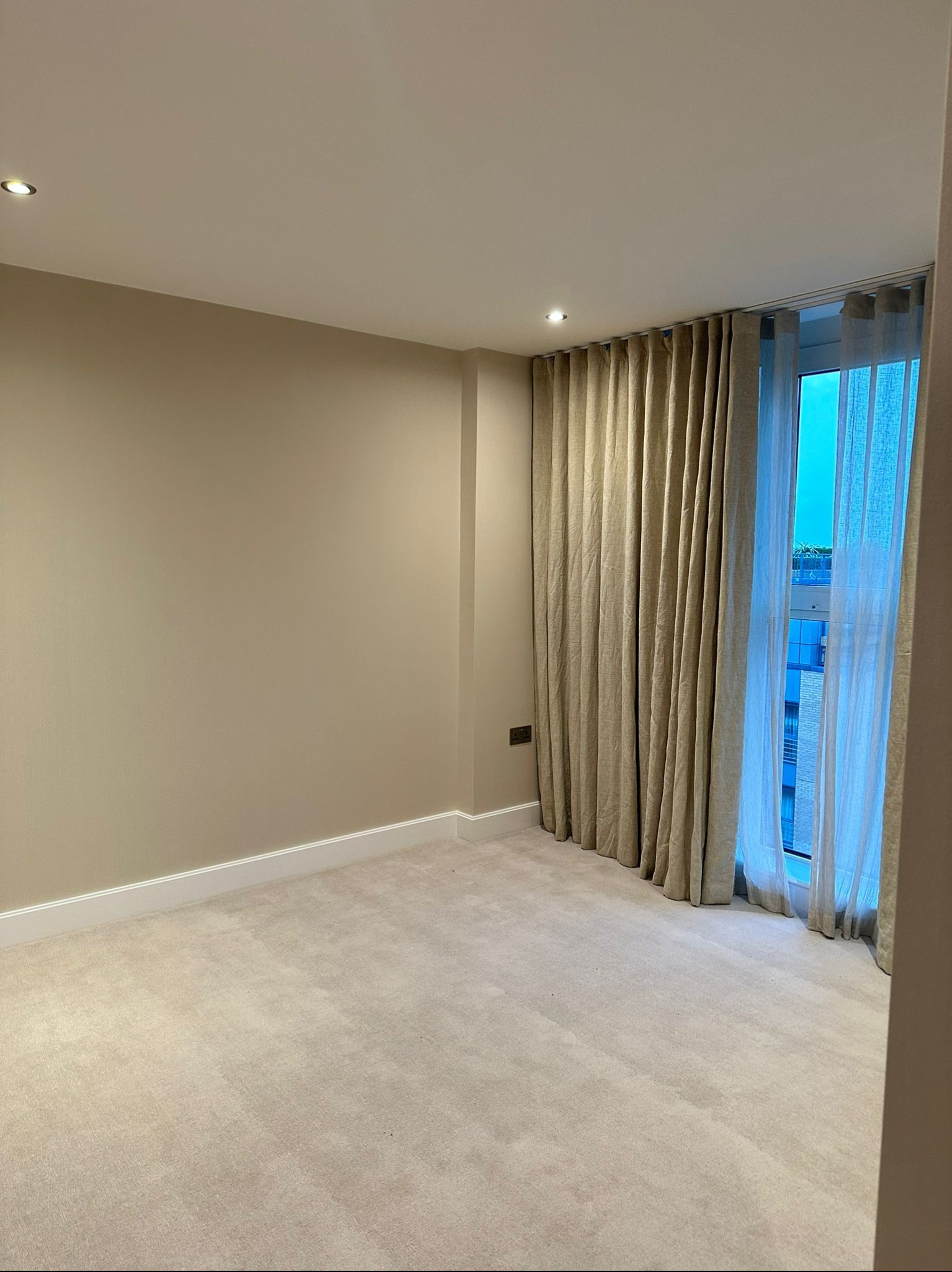 Pusty pokój z beżowymi ścianami, jasną wykładziną dywanową i zasłonami w oknie, widoczny fragment budynku za oknem.