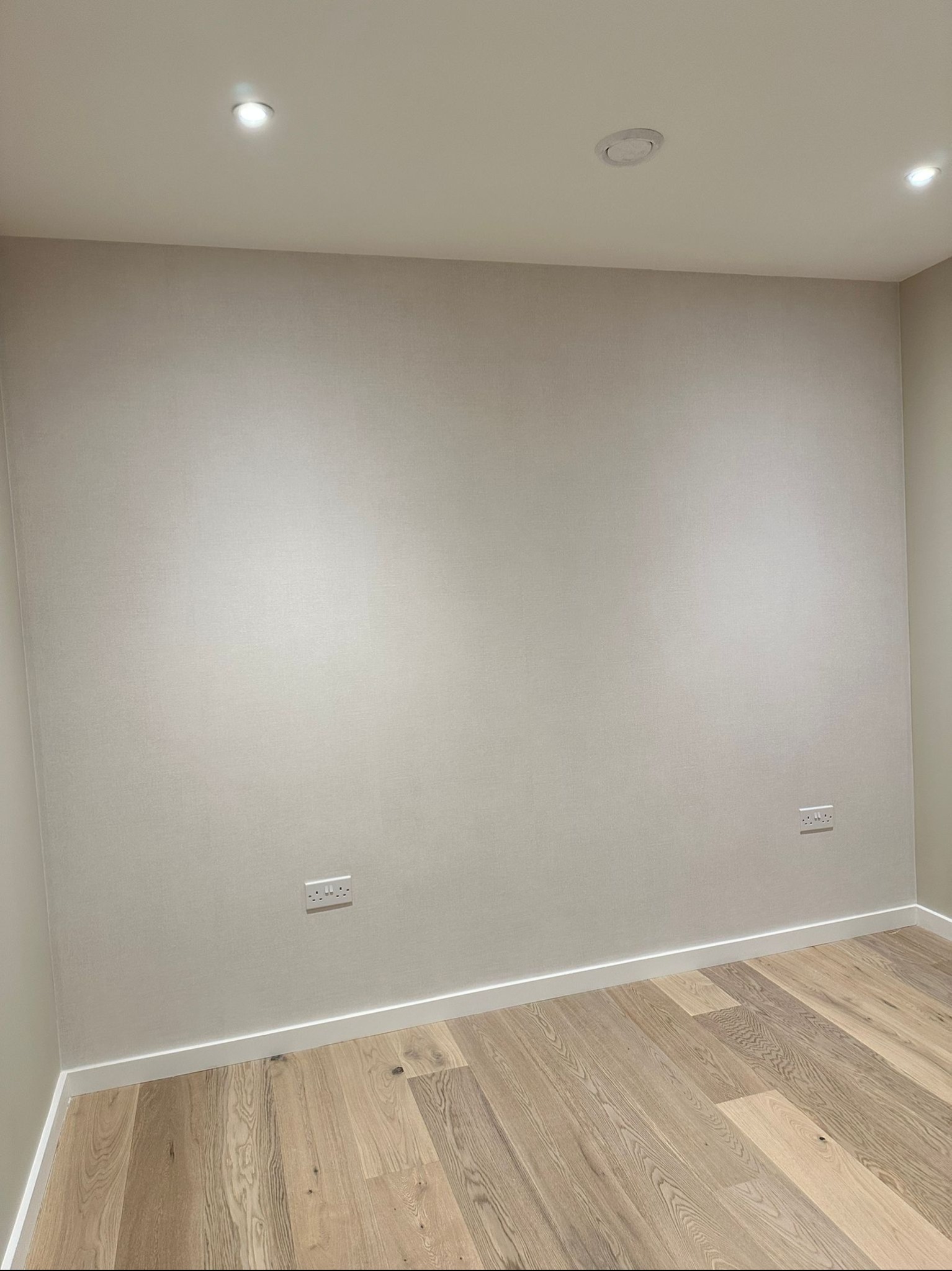 Pomalowane na jasny odcień ściany w pokoju z drewnianą podłogą i listwami przypodłogowymi, widoczne gniazdka elektryczne i oświetlenie sufitowe.