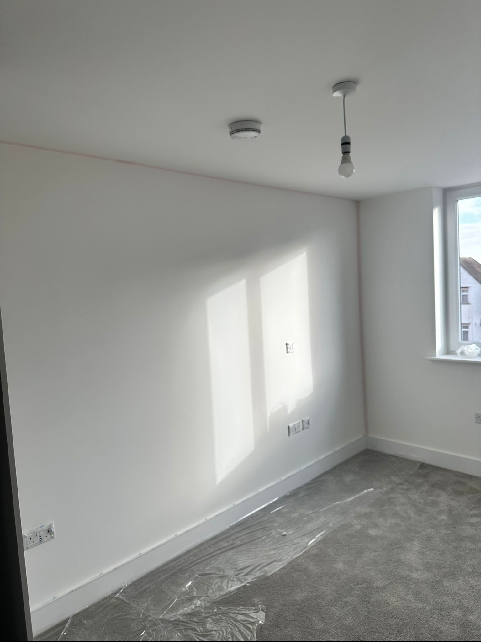 Pomalowane na biało wnętrze pokoju z szarym dywanem zabezpieczonym folią, widoczne gniazdka elektryczne, okno z widokiem na budynek i wisząca żarówka.