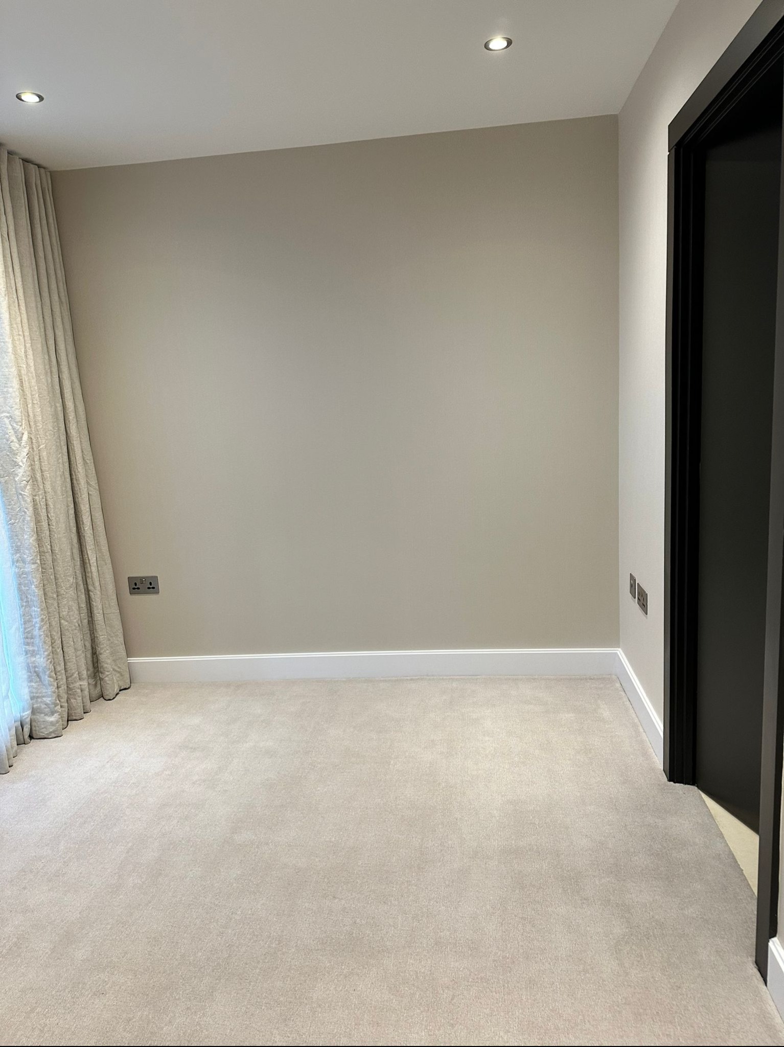 Pusty pokój z beżową ścianą, kremowym dywanem, białymi listwami przypodłogowymi i czarnymi drzwiami, oświetlony punktowo.