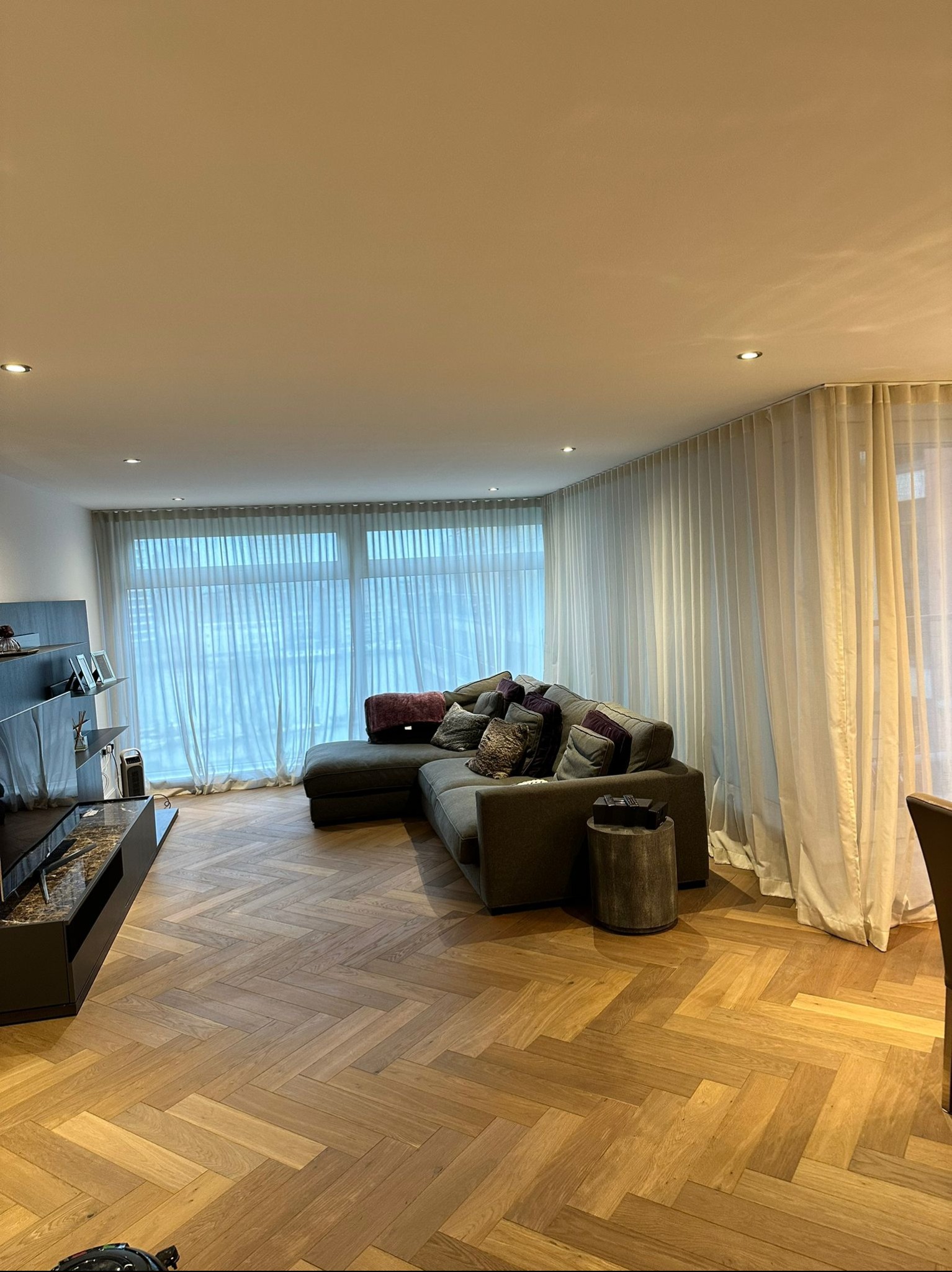 Salon z parkietem ułożonym w jodełkę, szarą sofą narożną i białymi, zwiewnymi zasłonami sięgającymi od sufitu do podłogi.