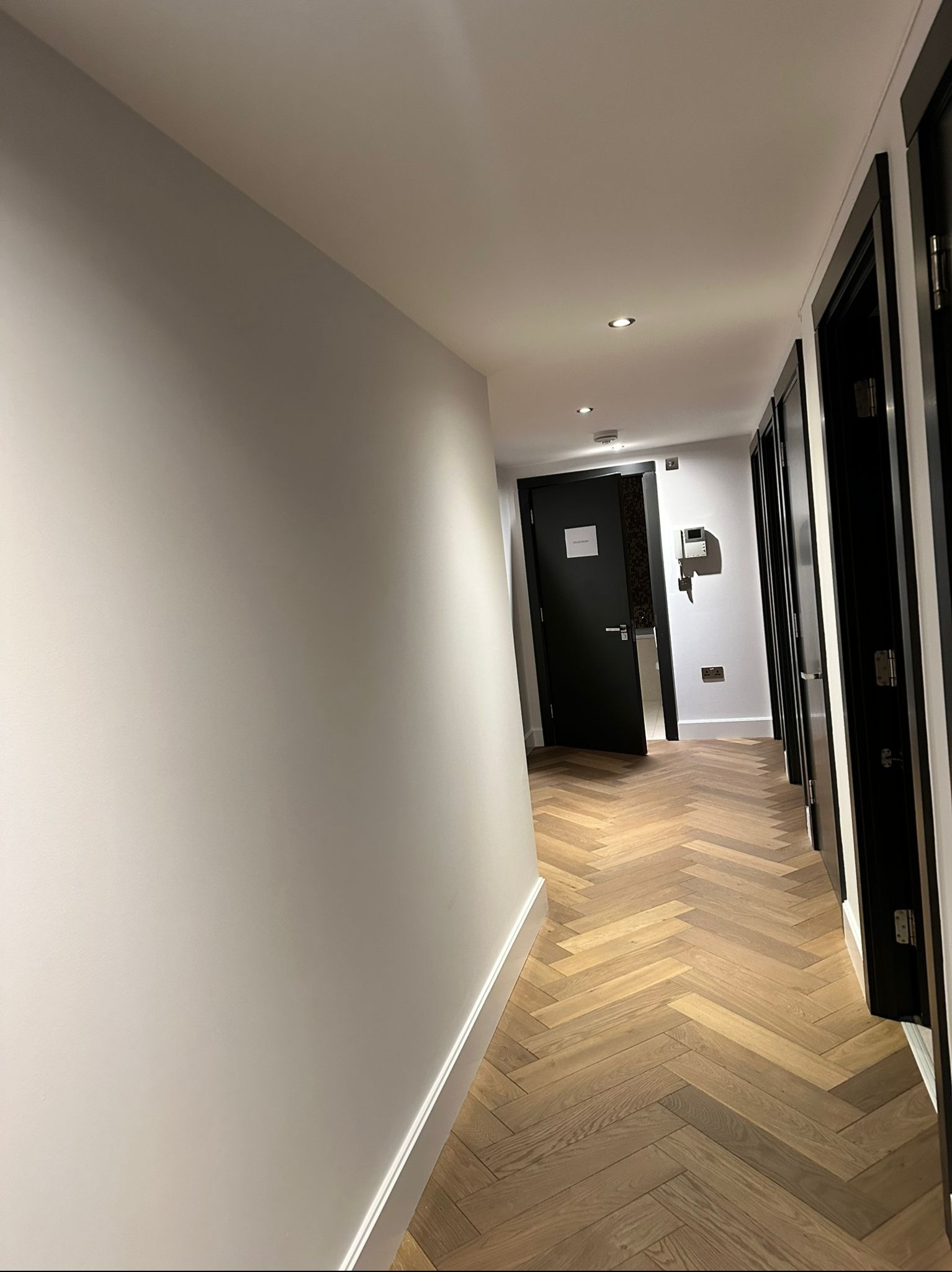 Korytarz z parkietem ułożonym w jodełkę, białe ściany i czarne drzwi, widok w perspektywie.