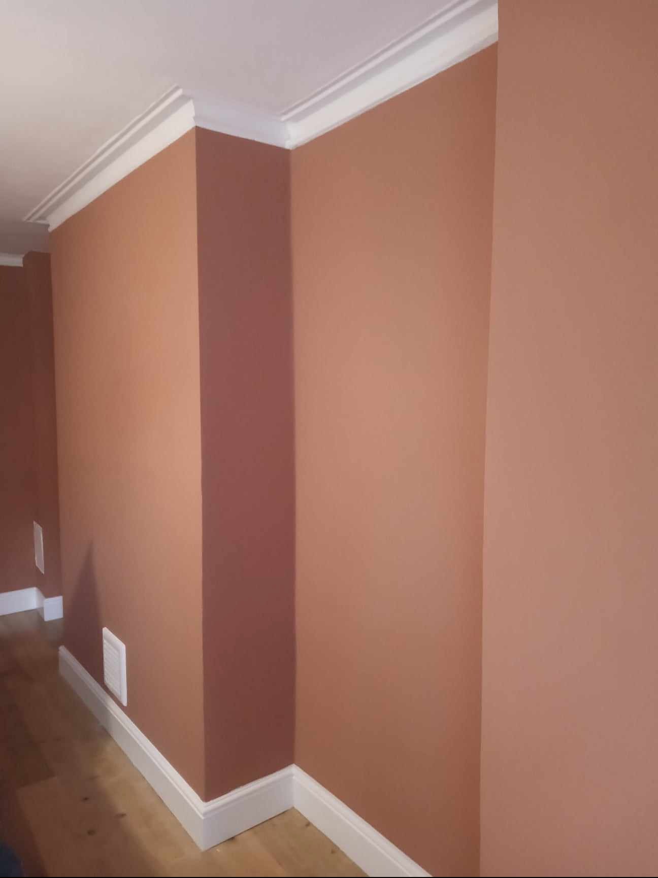 Pomarańczowo-brązowe ściany po świeżym malowaniu z białymi listwami przypodłogowymi i sufitowymi, widoczne kratki wentylacyjne.