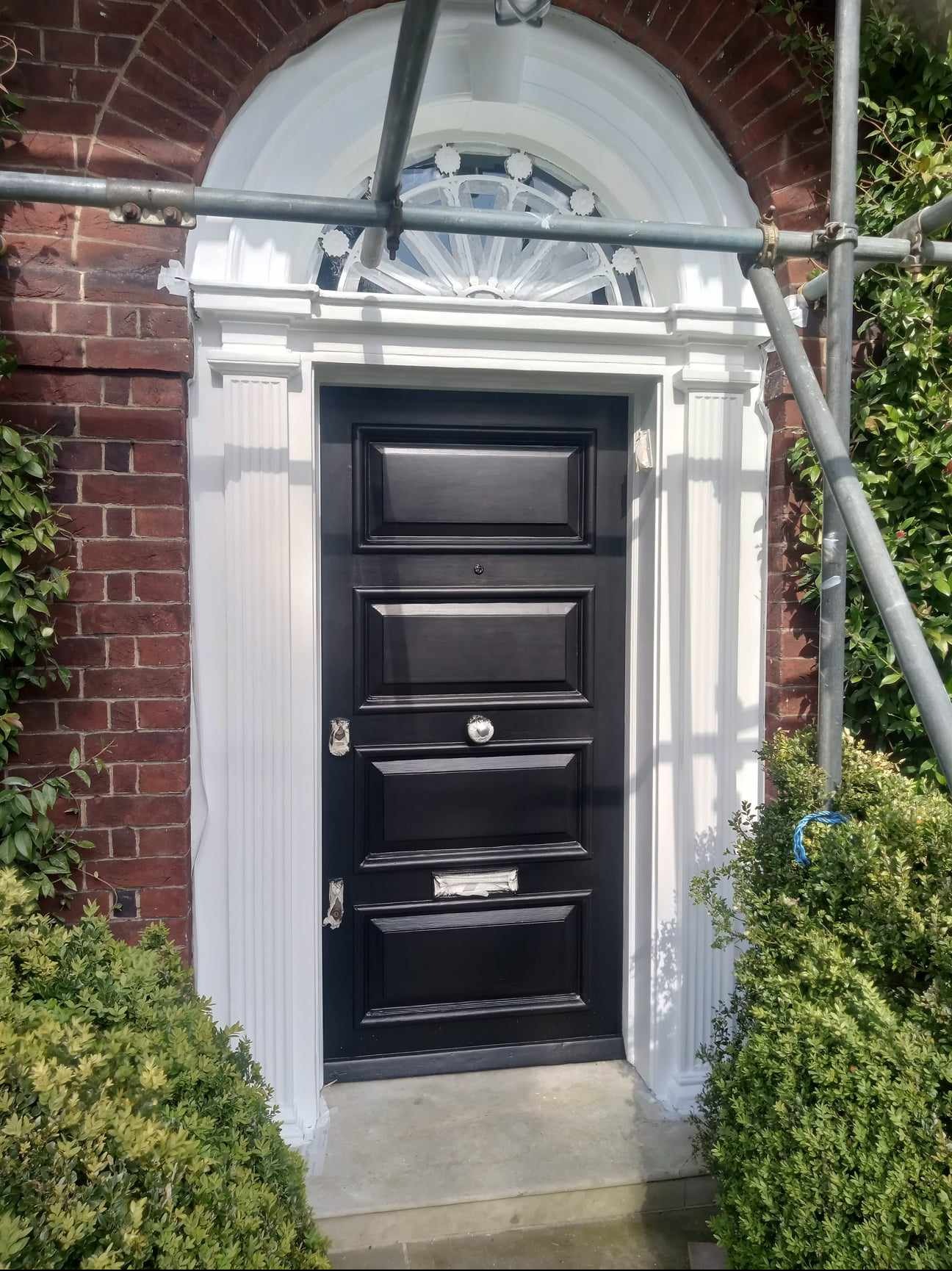 Czarna, panelowa brama wejściowa w trakcie malowania ościeżnicy na biało, widoczne rusztowanie i ceglana elewacja.