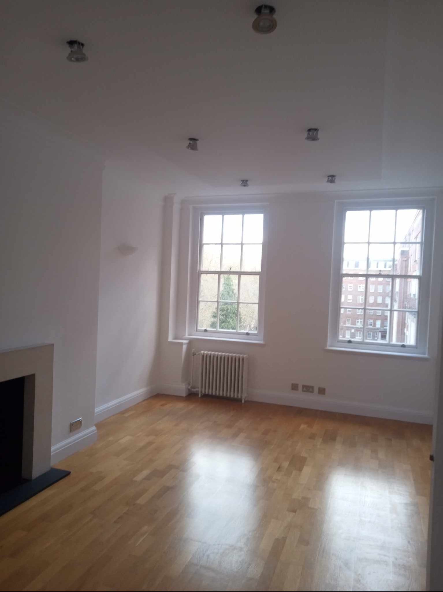 Pomalowane na biało wnętrze pokoju z parkietem, grzejnikiem pod oknem i kominkiem po lewej stronie.