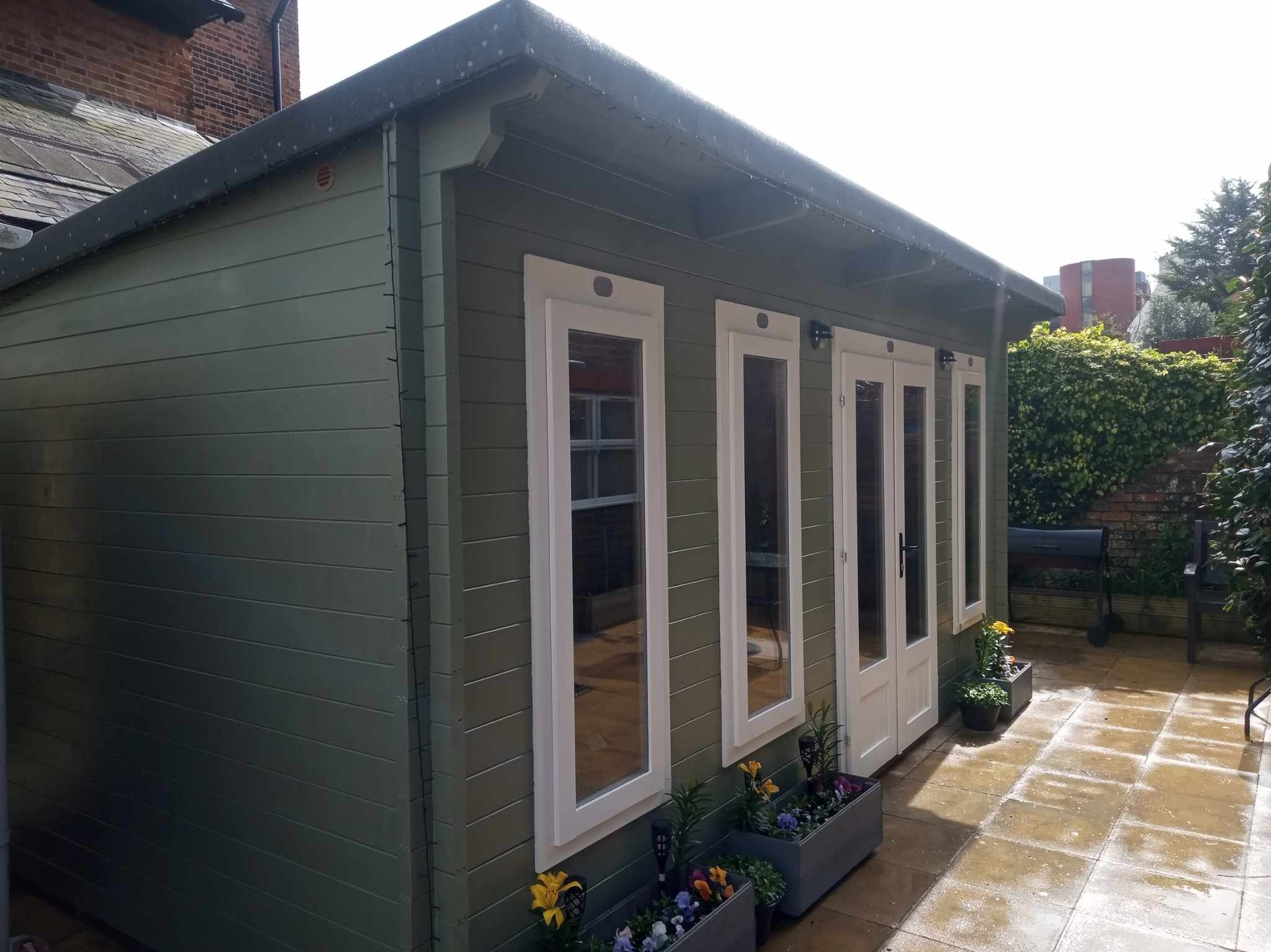 Nowoczesny, zielony domek ogrodowy z białymi ramami okiennymi i przeszklonymi drzwiami, otoczony kwiatami w donicach na mokrym, wyłożonym płytkami patio.