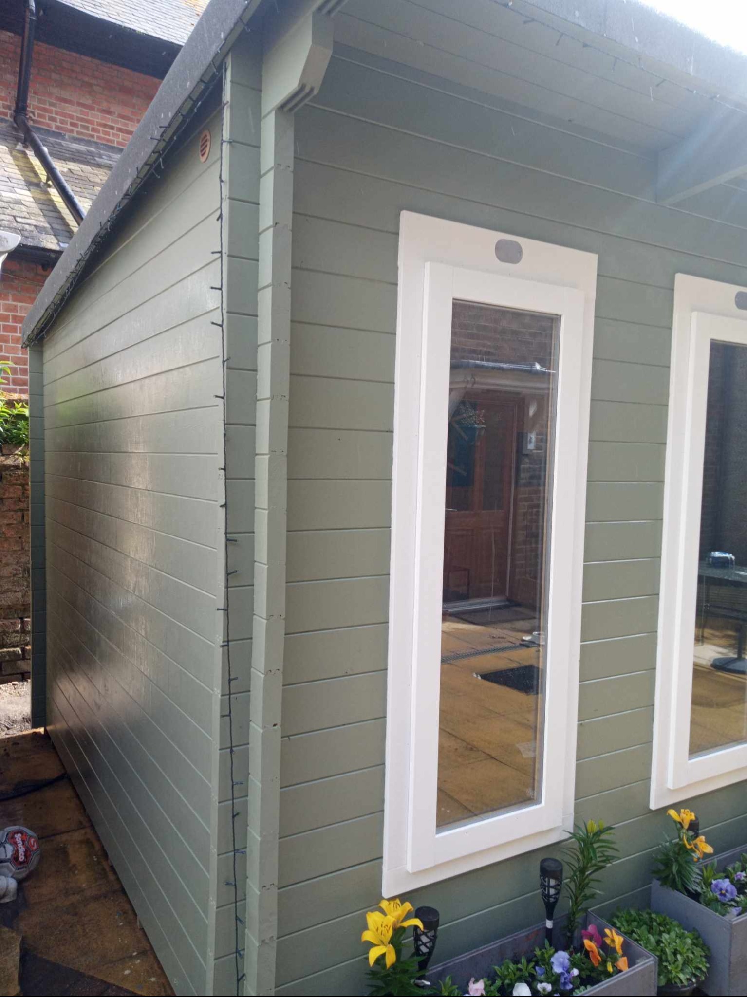 Świeżo pomalowany, drewniany domek ogrodowy w odcieniach zieleni z białymi ramami okiennymi, otoczony rabatami kwiatowymi, odbicie domu w szybach.