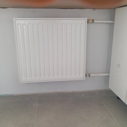 Hydroinstalmonter - Biały, panelowy grzejnik zamontowany na szarej ścianie, z widocznymi rurami doprowadzającymi i zaworami, obok biała szafka.