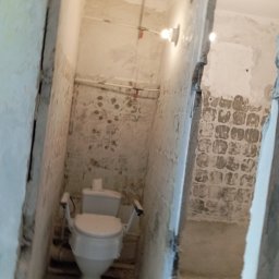 Hydroinstalmonter - Wnętrze łazienki w trakcie remontu z widoczną toaletą z uchwytami dla osób niepełnosprawnych, rurami biegnącymi wzdłuż ściany i surowymi ścianami bez płytek.