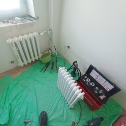 Hydroinstalmonter - Dwa białe grzejniki żeberkowe, jeden zamontowany przy oknie, drugi leżący na zielonej folii ochronnej, obok skrzynka z narzędziami, wiertarka i torba narzędziowa w trakcie prac instalacyjnych...