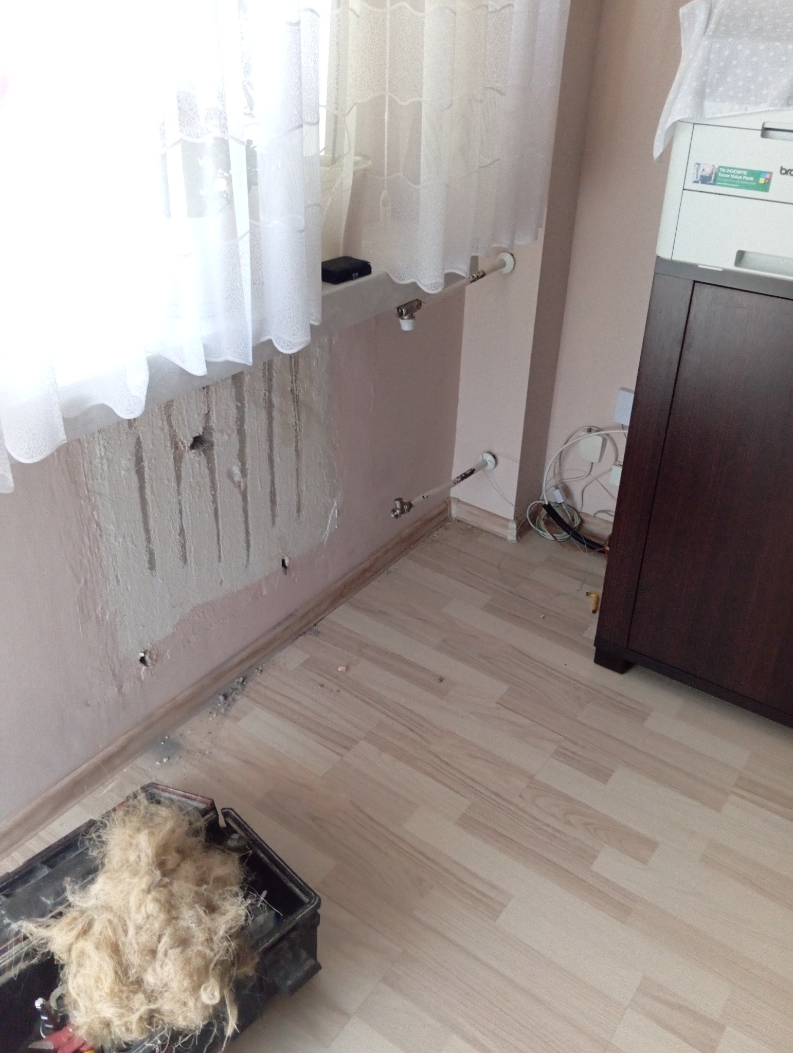 Ściana po demontażu grzejnika z widocznymi śladami po rurach, otwory w ścianie, skrzynka z wełną mineralną na podłodze, fragment grzejnika przy oknie.