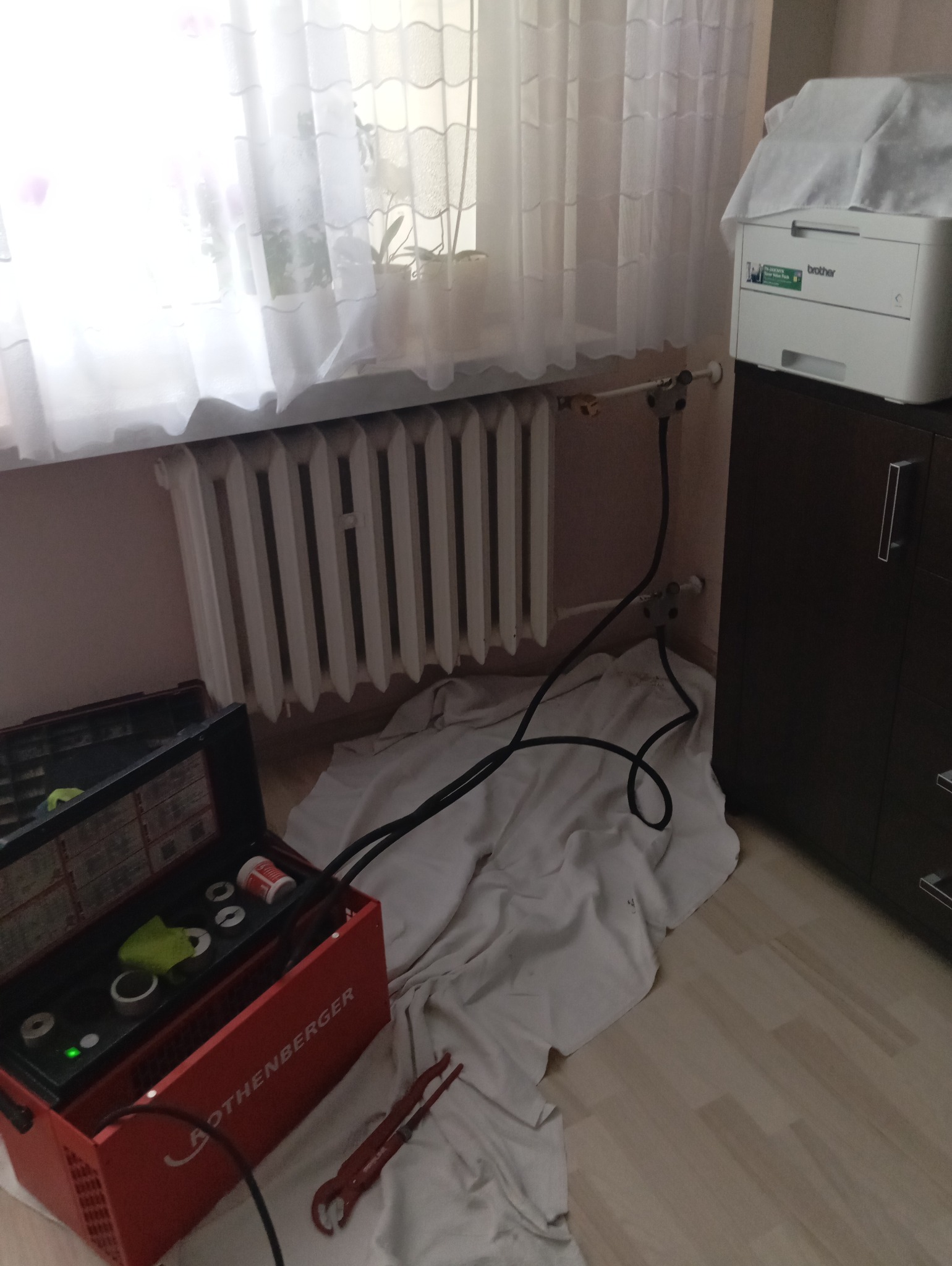 Podłączenie grzejnika do instalacji, widoczny grzejnik żeberkowy, rury instalacyjne, narzędzia Rothenberger i zabezpieczająca płachta na podłodze.