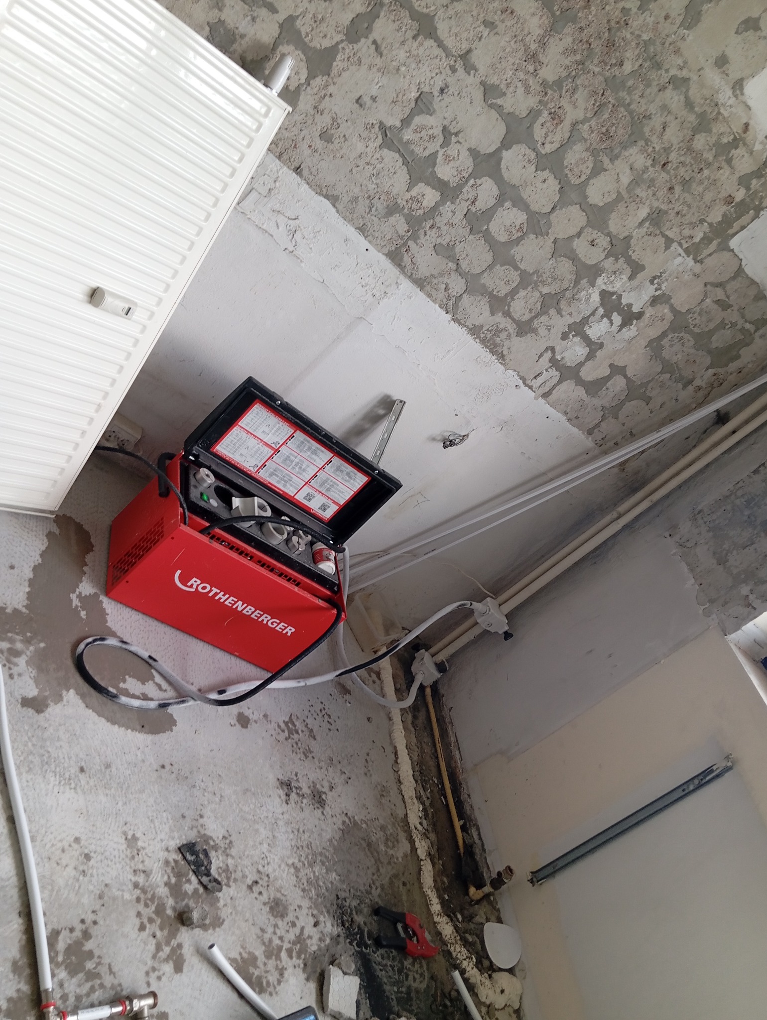 Czerwona zgrzewarka Rothenberger na podłodze w trakcie prac instalacyjnych, widoczne rury, grzejnik i surowy stan wykończenia pomieszczenia.