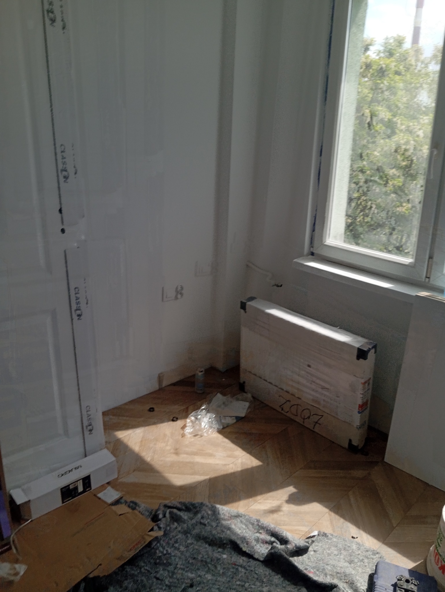 Rozpakowany grzejnik w folii ochronnej, leżący na podłodze w rogu pokoju, obok okna i drzwi, widoczne ślady po instalacji na ścianie.