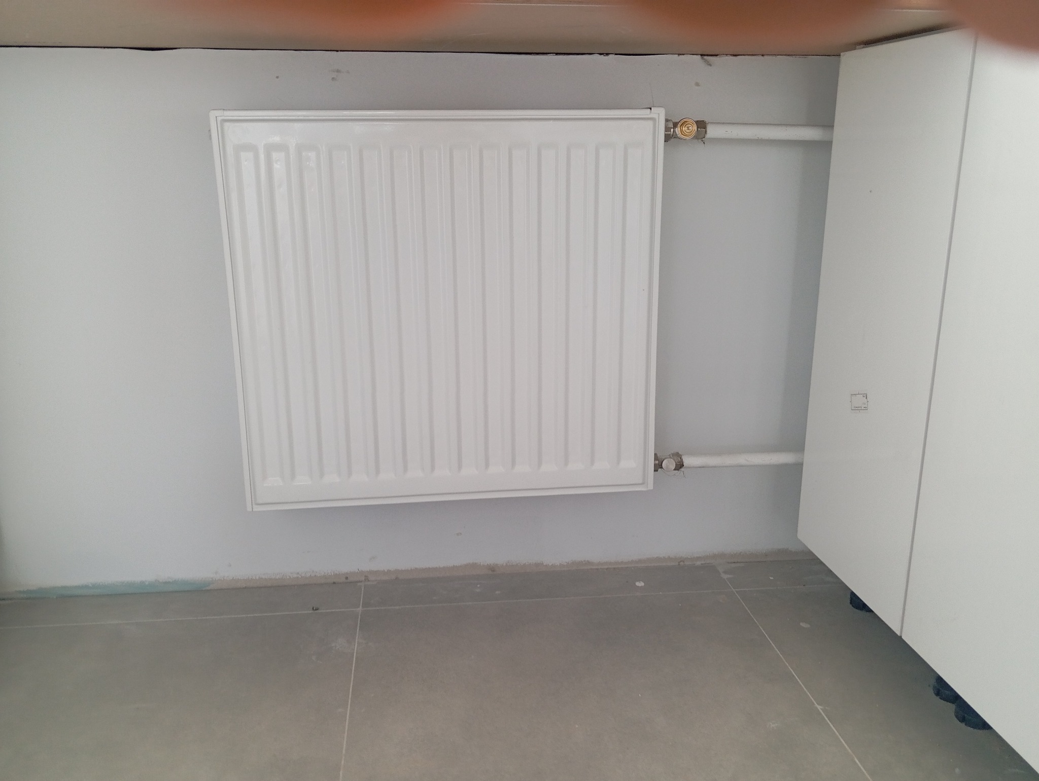 Biały, panelowy grzejnik zamontowany na szarej ścianie, z widocznymi rurami doprowadzającymi i zaworami, obok biała szafka.