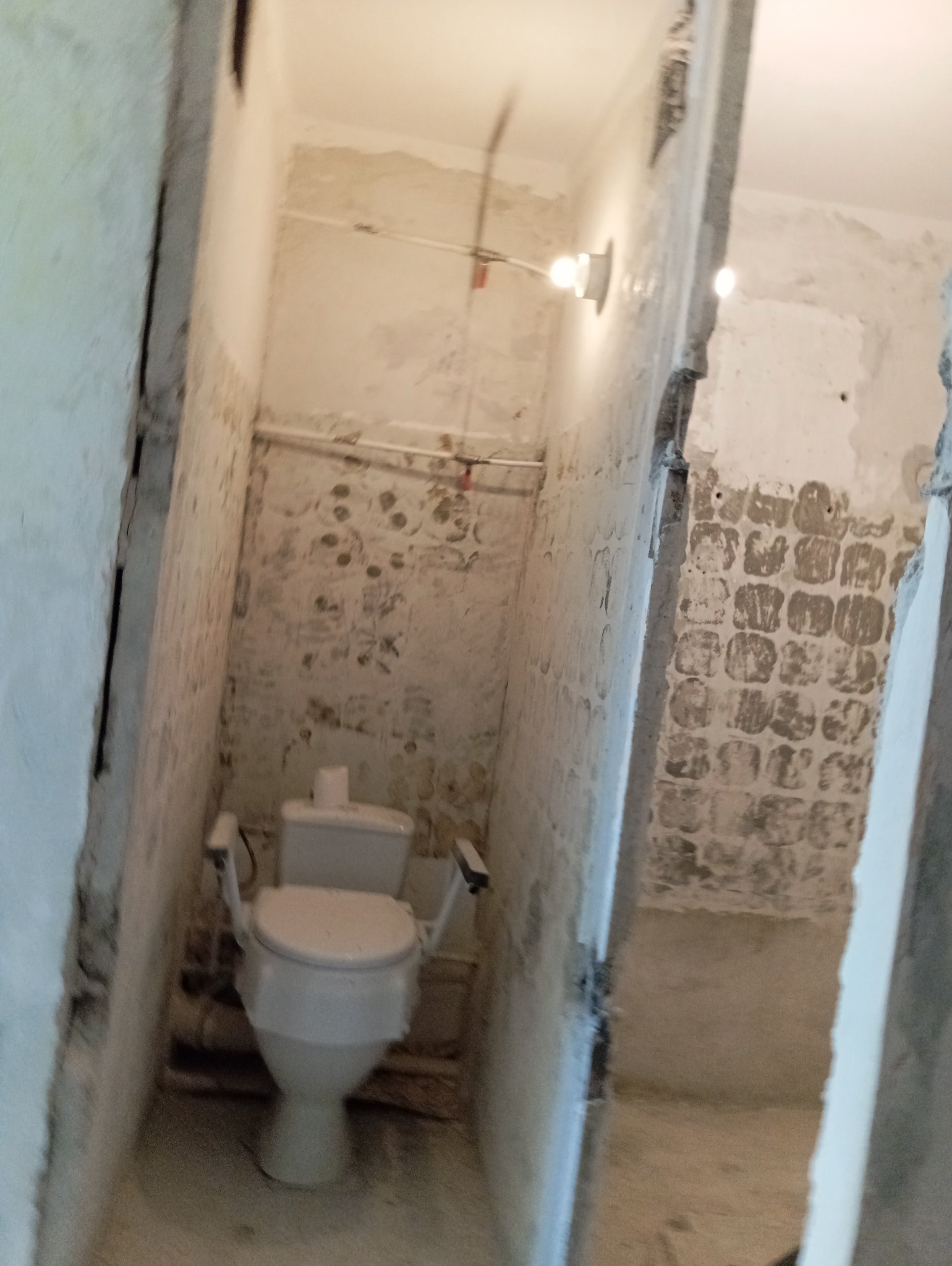 Wnętrze łazienki w trakcie remontu z widoczną toaletą z uchwytami dla osób niepełnosprawnych, rurami biegnącymi wzdłuż ściany i surowymi ścianami bez płytek.