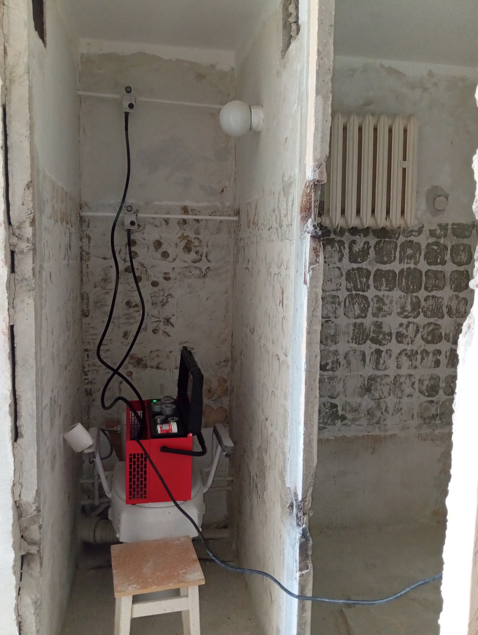 Czerwone urządzenie diagnostyczne podłączone do instalacji w trakcie remontu łazienki, widoczne ślady po starych płytkach i grzejnik żeberkowy w tle.