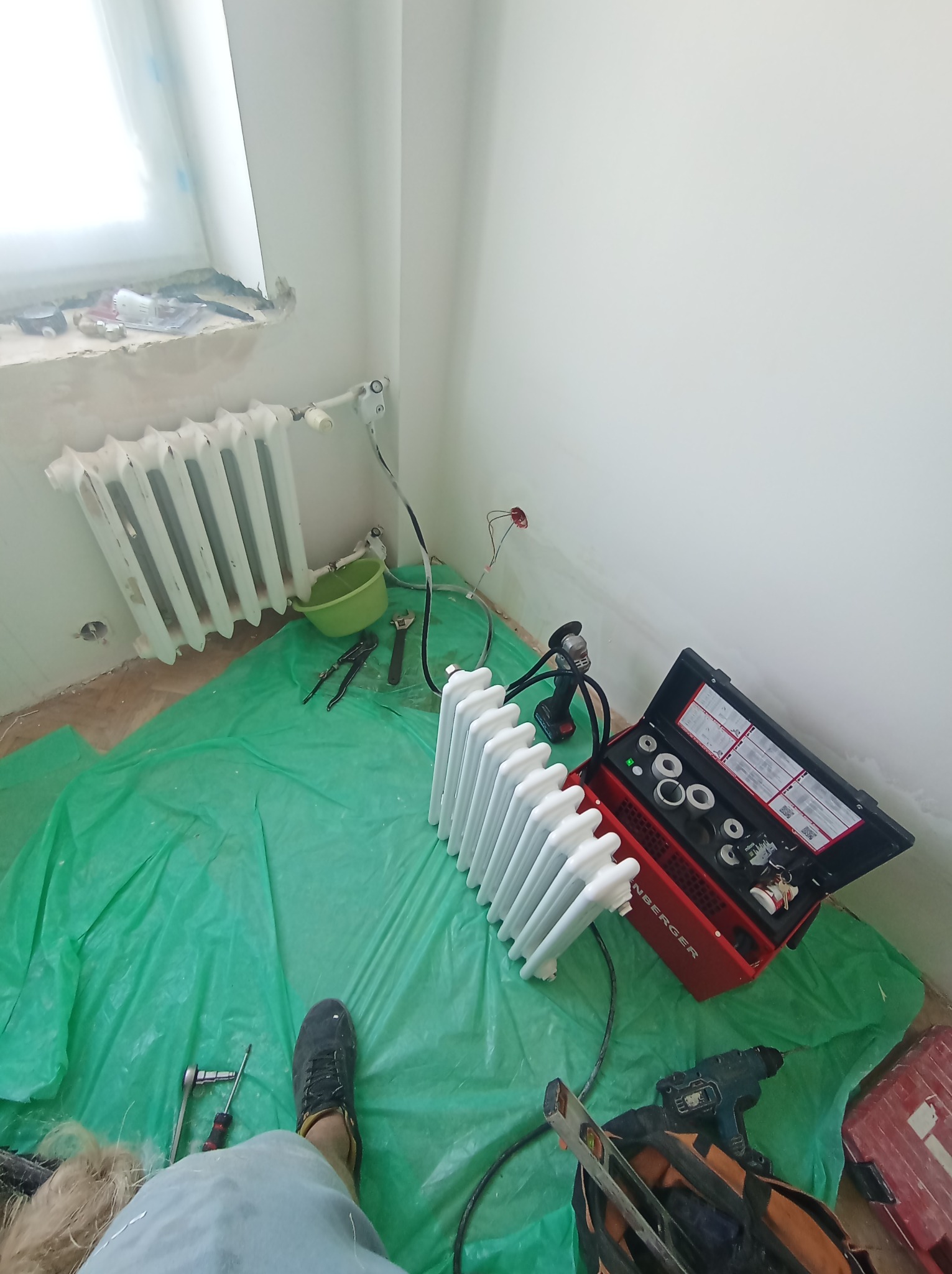 Dwa białe grzejniki żeberkowe, jeden zamontowany przy oknie, drugi leżący na zielonej folii ochronnej, obok skrzynka z narzędziami, wiertarka i torba narzędziowa w trakcie prac instalacyjnych...
