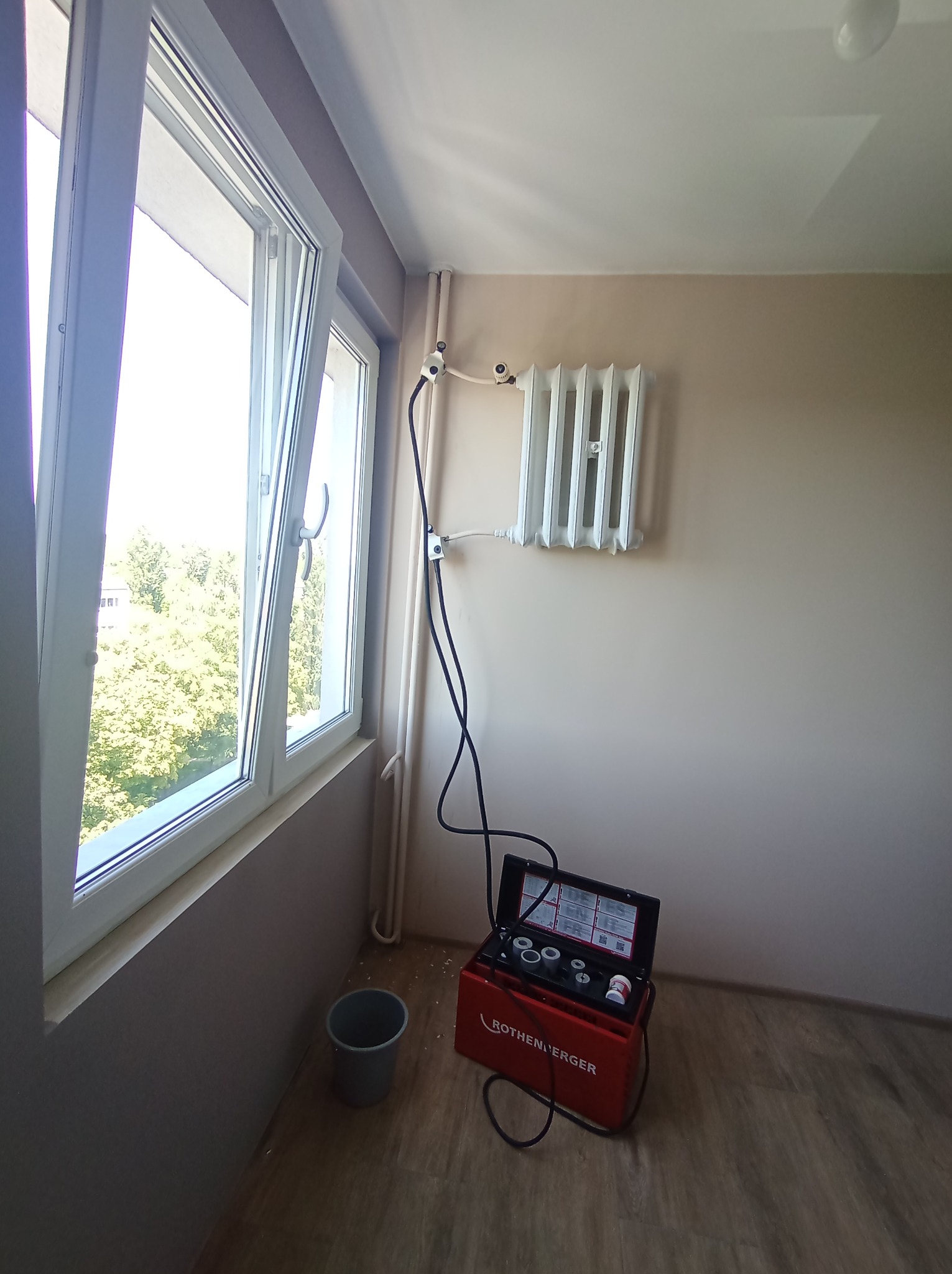 Biały, żeliwny grzejnik panelowy zamontowany na beżowej ścianie, podłączony do instalacji. Obok okna otwartego na oścież stoi czerwona walizka z narzędziami firmy Rothenberger i szary kubełek.