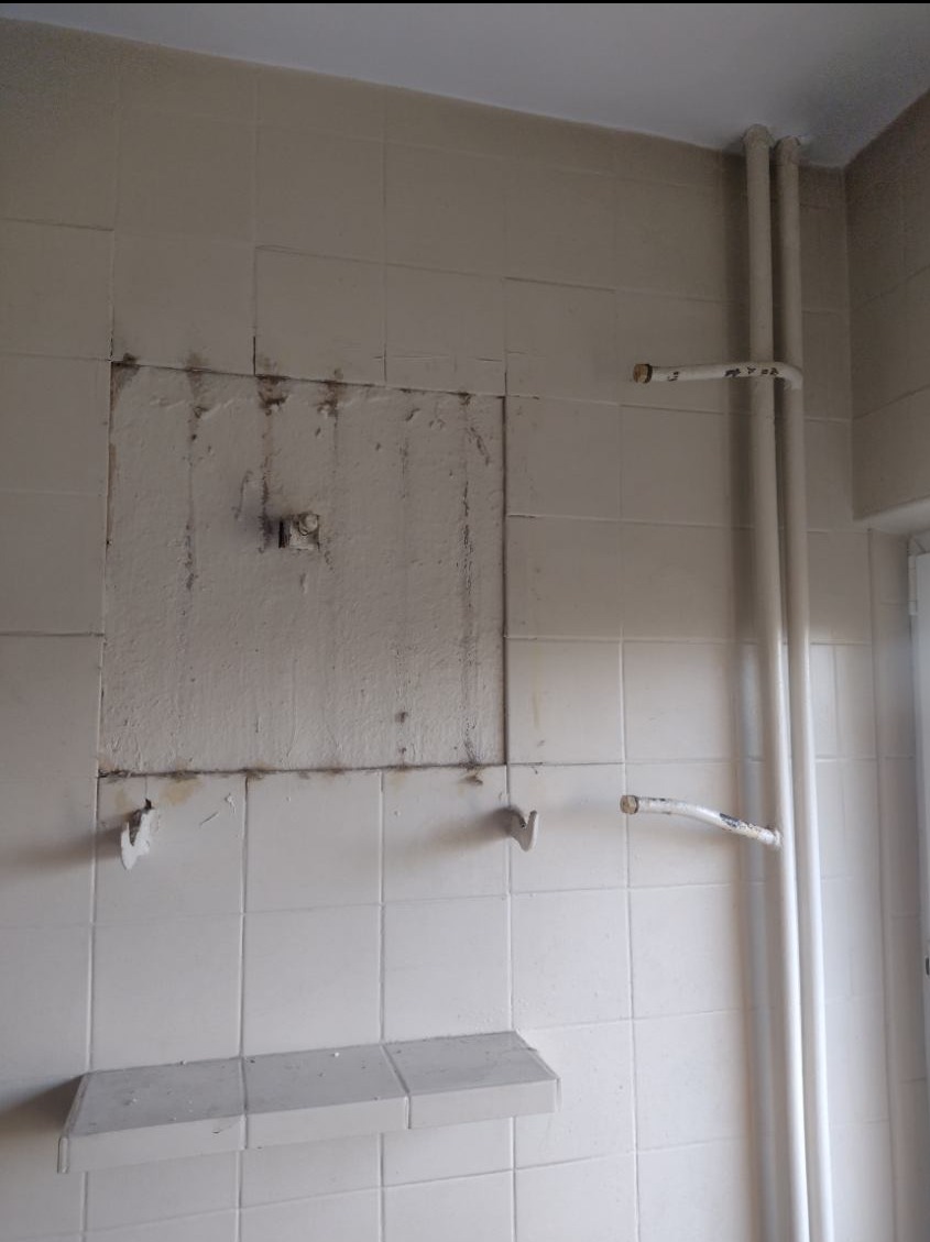 Ściana w łazience z białymi płytkami, fragment ściany z usuniętymi płytkami i odsłoniętą instalacją hydrauliczną, widoczne rury i uchwyty, półka z płytek, ślady po usuniętych płytkach.