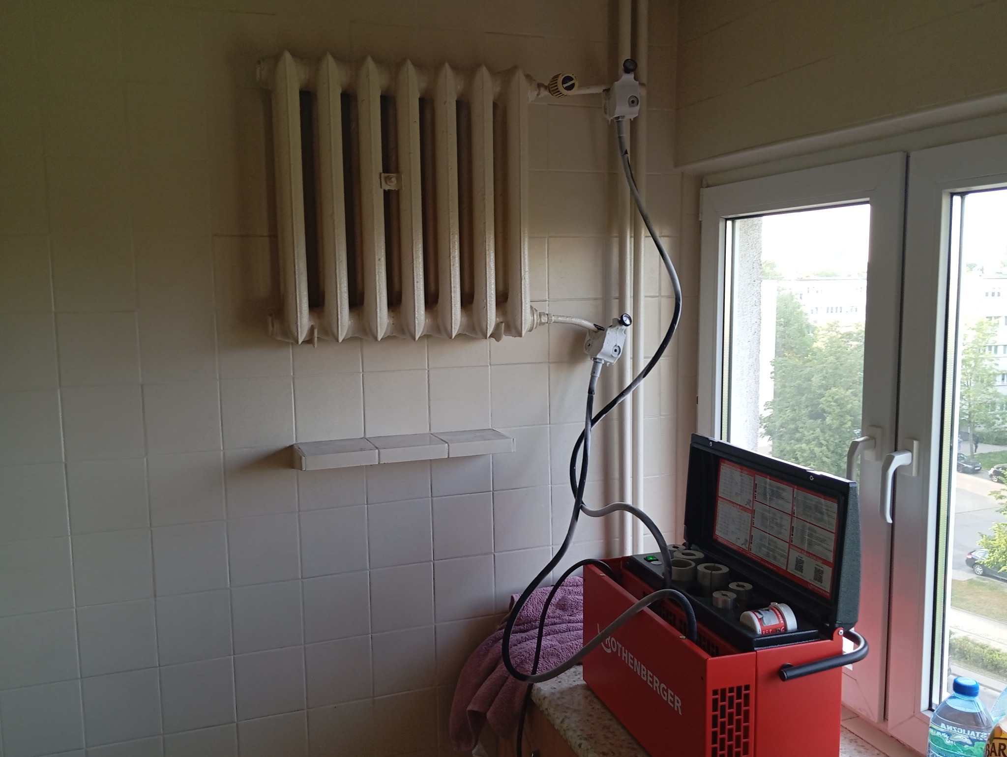 Staromodny, żeliwny grzejnik na ścianie z białych płytek, podłączony do urządzenia pomiarowego Rothenberger, stojącego na szafce przy oknie.