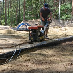 RAF usługi budowlane - Pracownik w pomarańczowych rękawicach zagęszcza piasek zagęszczarką wibracyjną Ramirent, widoczne rury i czarna folia izolacyjna fundamentów, w tle las iglasty.