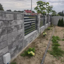 Nowoczesne ogrodzenie z szarego betonu z poziomymi, metalowymi przęsłami, oddzielające posesję. Widoczne młode drzewka i krzewy hortensji wzdłuż ogrodzenia.