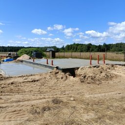 RAF usługi budowlane - Wylany fundament pod dom z wystającymi rurami wentylacyjnymi na tle błękitnego nieba z chmurami. Widoczny piasek i pracownicy w oddali.