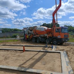 RAF usługi budowlane - Wylewanie betonu do szalunku fundamentów domu jednorodzinnego za pomocą pompy z betonomieszarki MAN. Na budowie dwóch pracowników w odzieży roboczej.