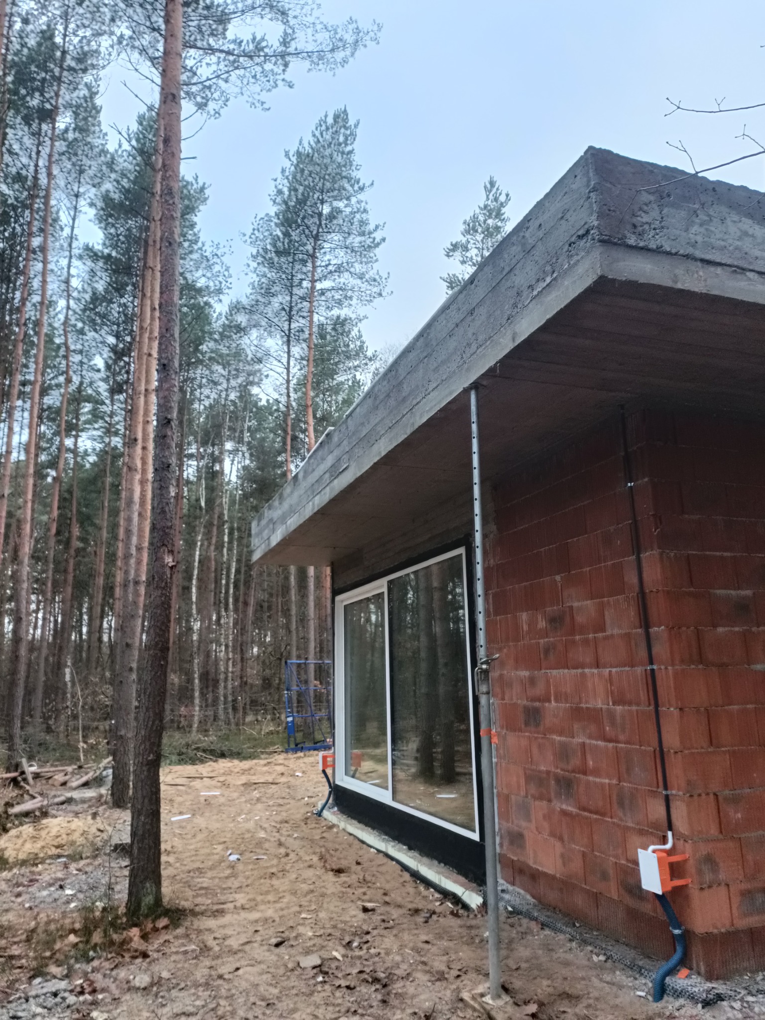 Surowy budynek z czerwonej cegły w trakcie budowy, widoczne okno, betonowy strop podparty metalowymi stemplami, instalacja elektryczna na zewnątrz, w tle las sosnowy i piasek.