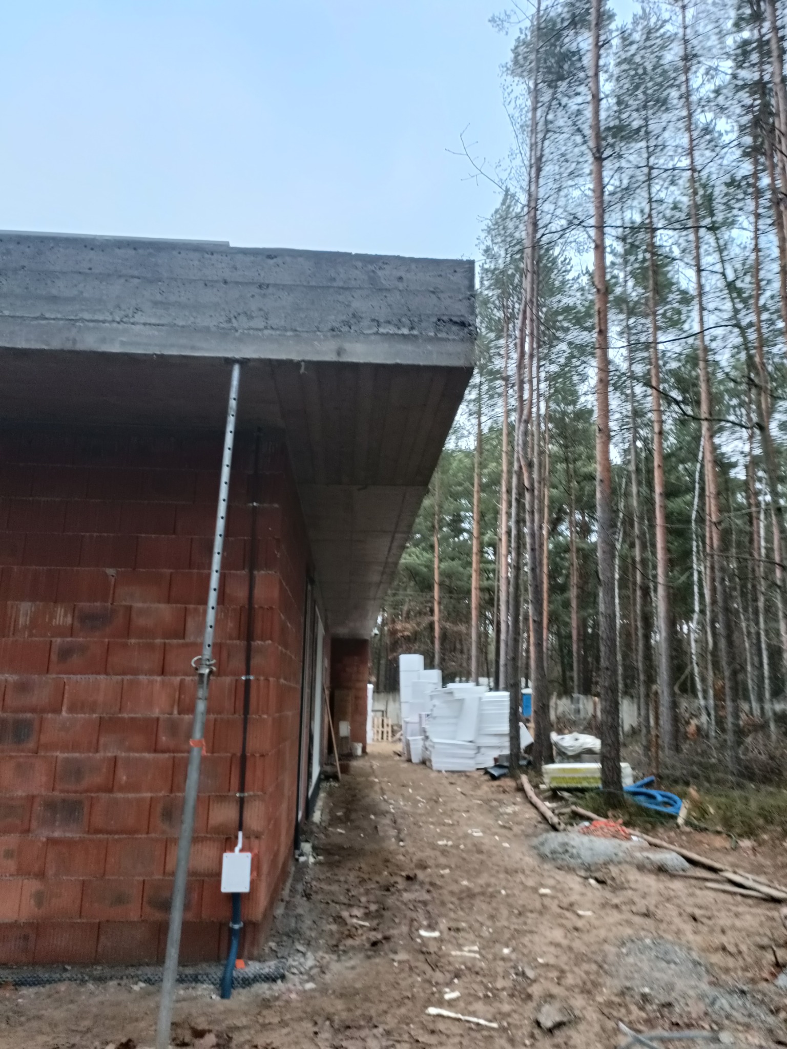 Surowy stan budynku z czerwonej cegły z betonowym stropodachem, podparty metalowym stemplem, widok z zewnątrz na tle lasu i sterty białego styropianu.
