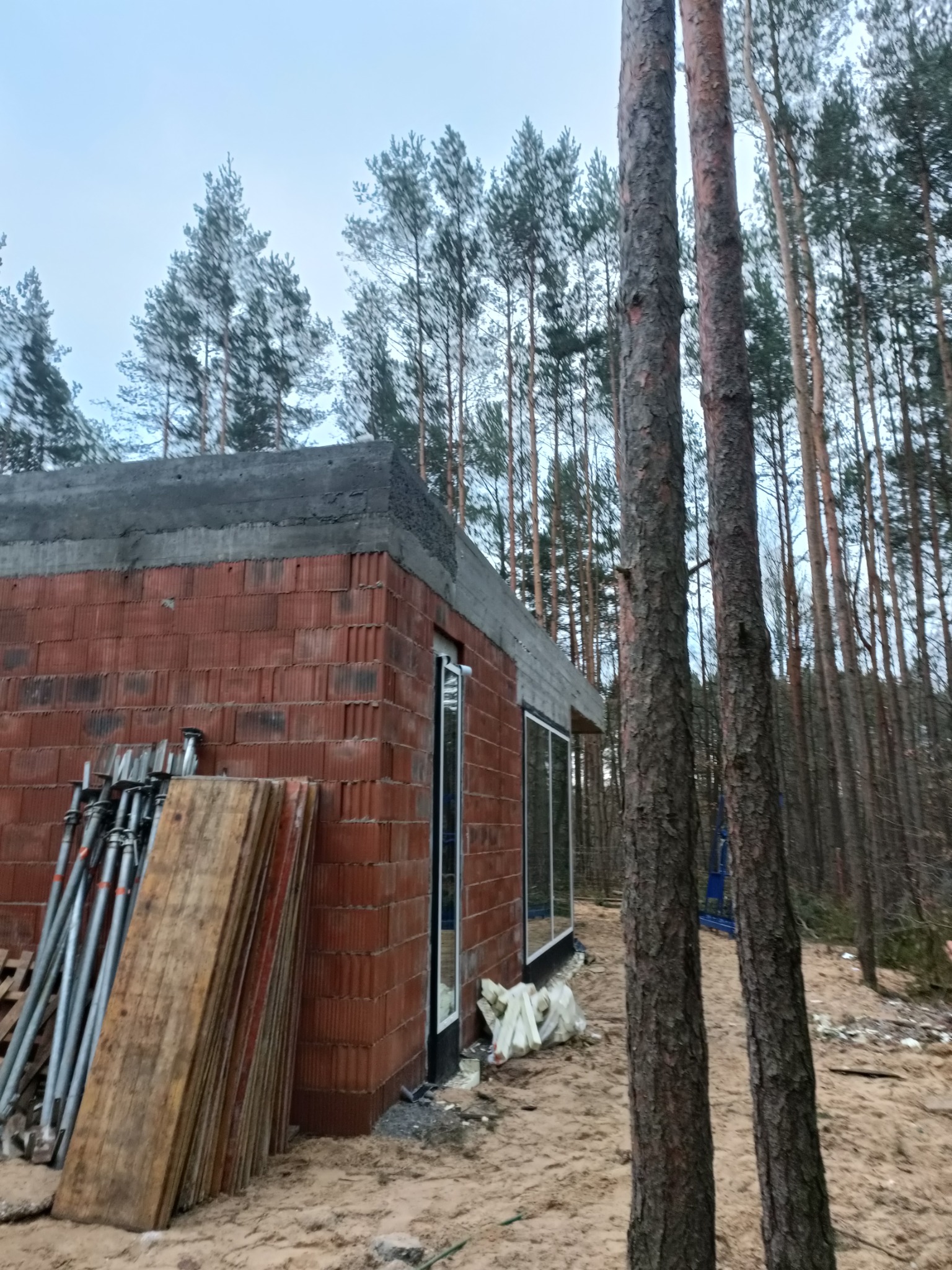 Surowy stan domu z czerwonej cegły, widoczne okna z białymi ramami, szalunki i podpory budowlane oparte o ścianę, w tle las iglasty i pochmurne niebo.