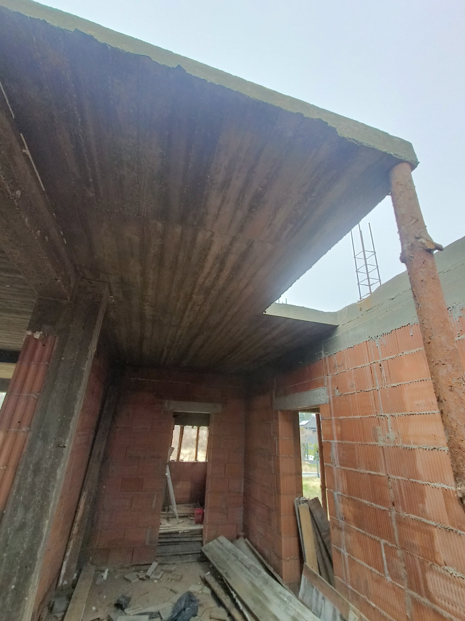 Wnętrze domu w trakcie budowy: ceglane ściany, betonowy strop z widocznym deskowaniem, surowe wykończenie i widok na zewnątrz przez okno.