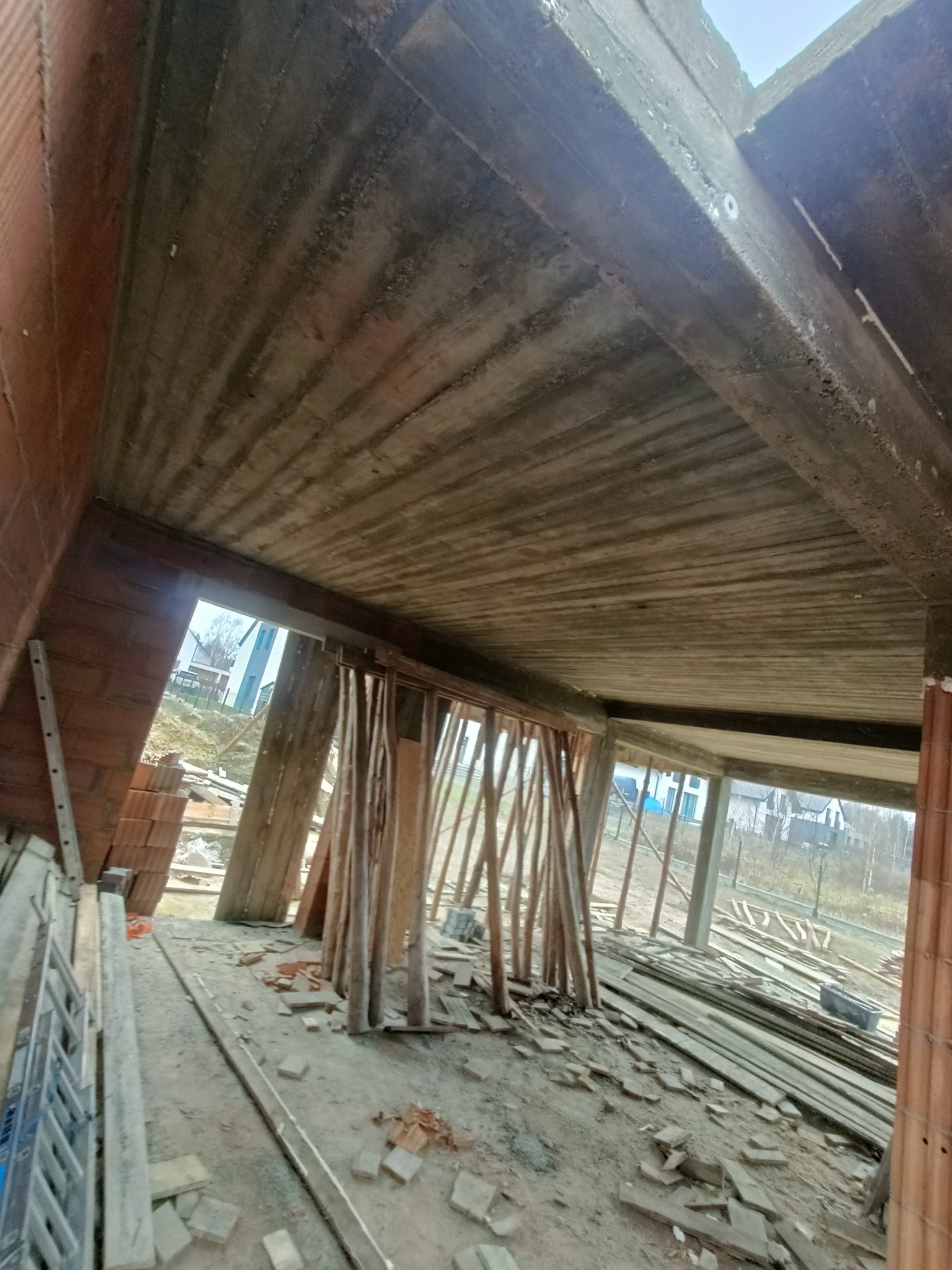 Wnętrze surowego domu w budowie: betonowy strop z drewnianymi podporami, fragmenty cegieł, widok na zewnątrz przez otwór okienny. W tle zabudowa jednorodzinna.