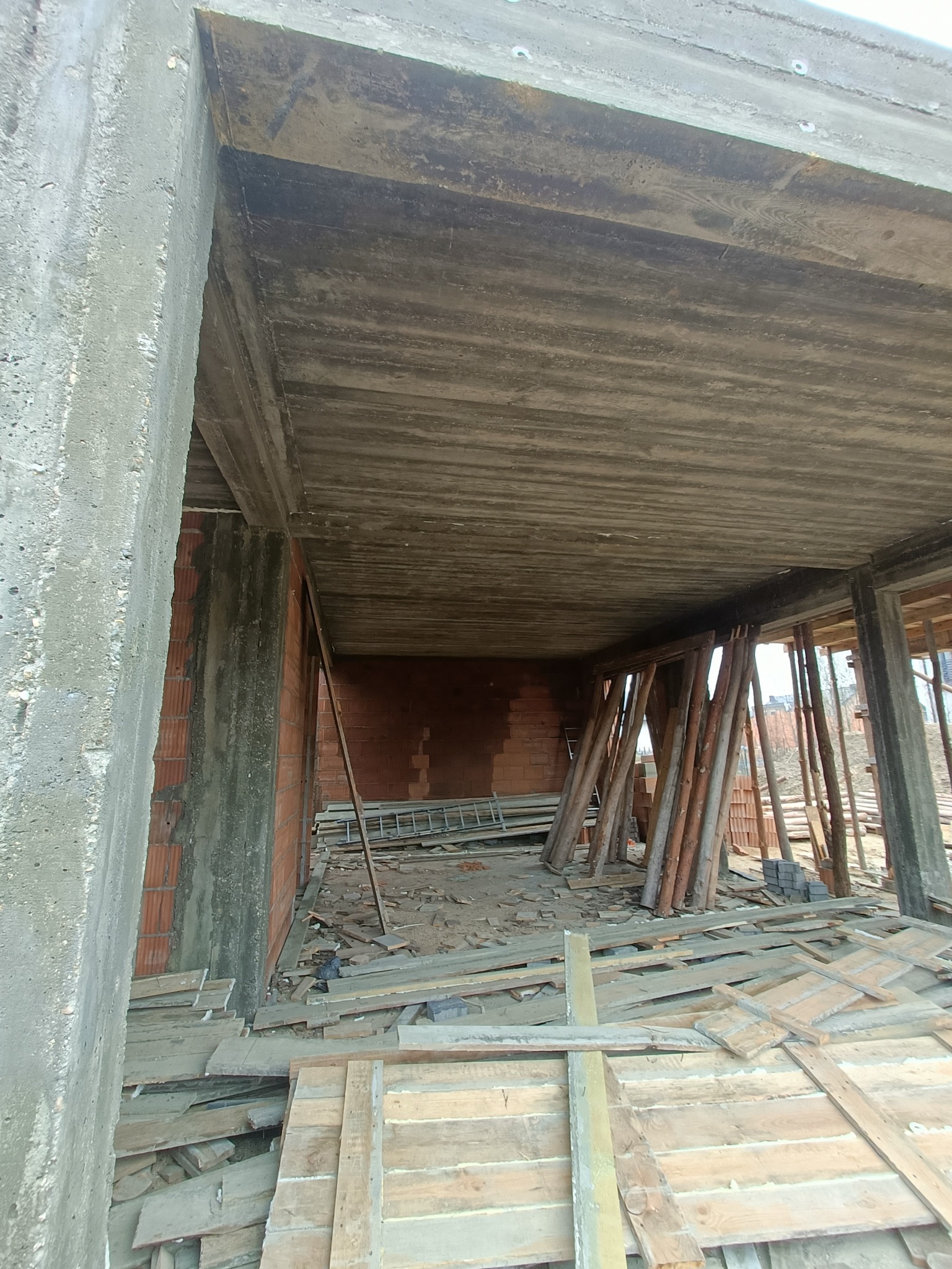 Surowy stan domu w budowie: betonowy strop z odciśniętym wzorem desek szalunkowych, ceglane ściany, drewniane stemple podtrzymujące strop, widok z wnętrza przez otwór drzwiowy.
