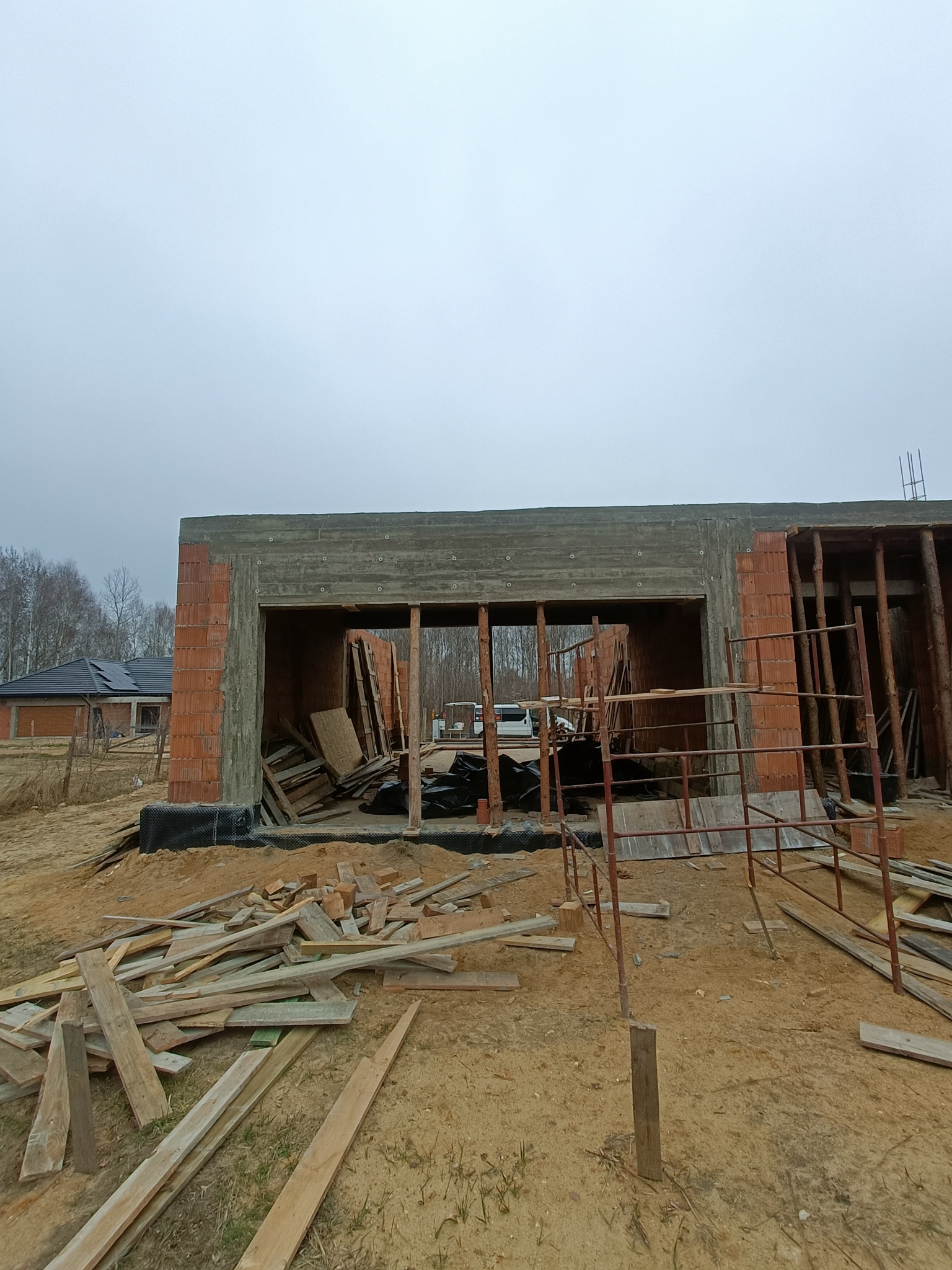 Budowa domu w stanie surowym otwartym: ceglane ściany, drewniane stemple, widoczne deski na ziemi, w tle budynek z czarnym dachem i szare niebo.