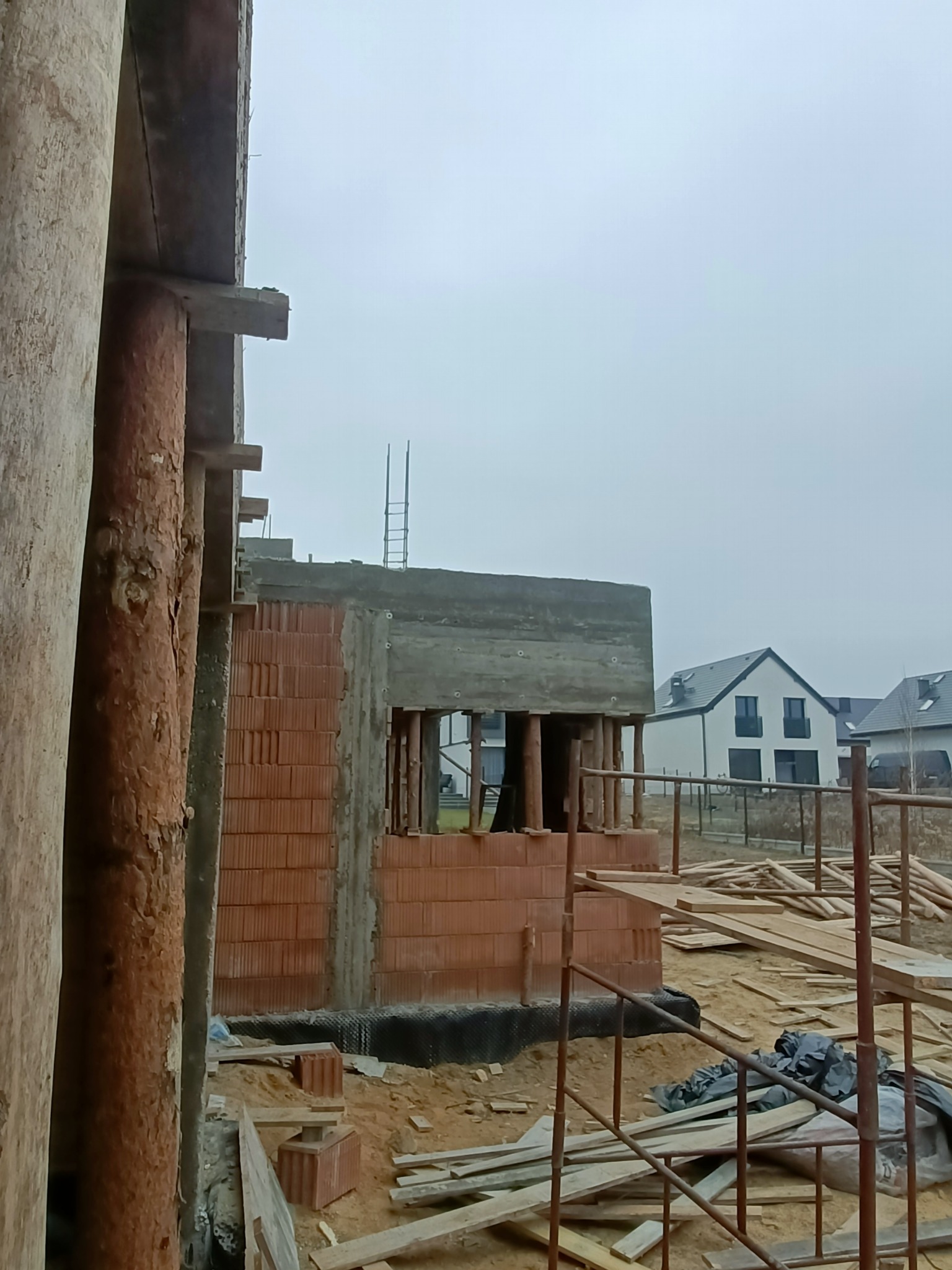 Surowy stan domu z czerwonej cegły, widok przez otwór w konstrukcji, drewniane podpory i deski na pierwszym planie, domy w tle, pochmurne niebo.