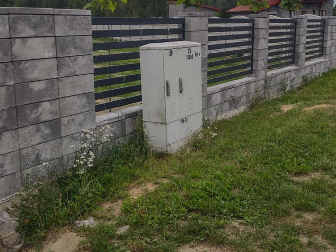 Szary, murowany płot z poziomymi, metalowymi przęsłami, z widoczną skrzynką elektryczną, otoczony zielenią. Ujęcie z poziomu gruntu.