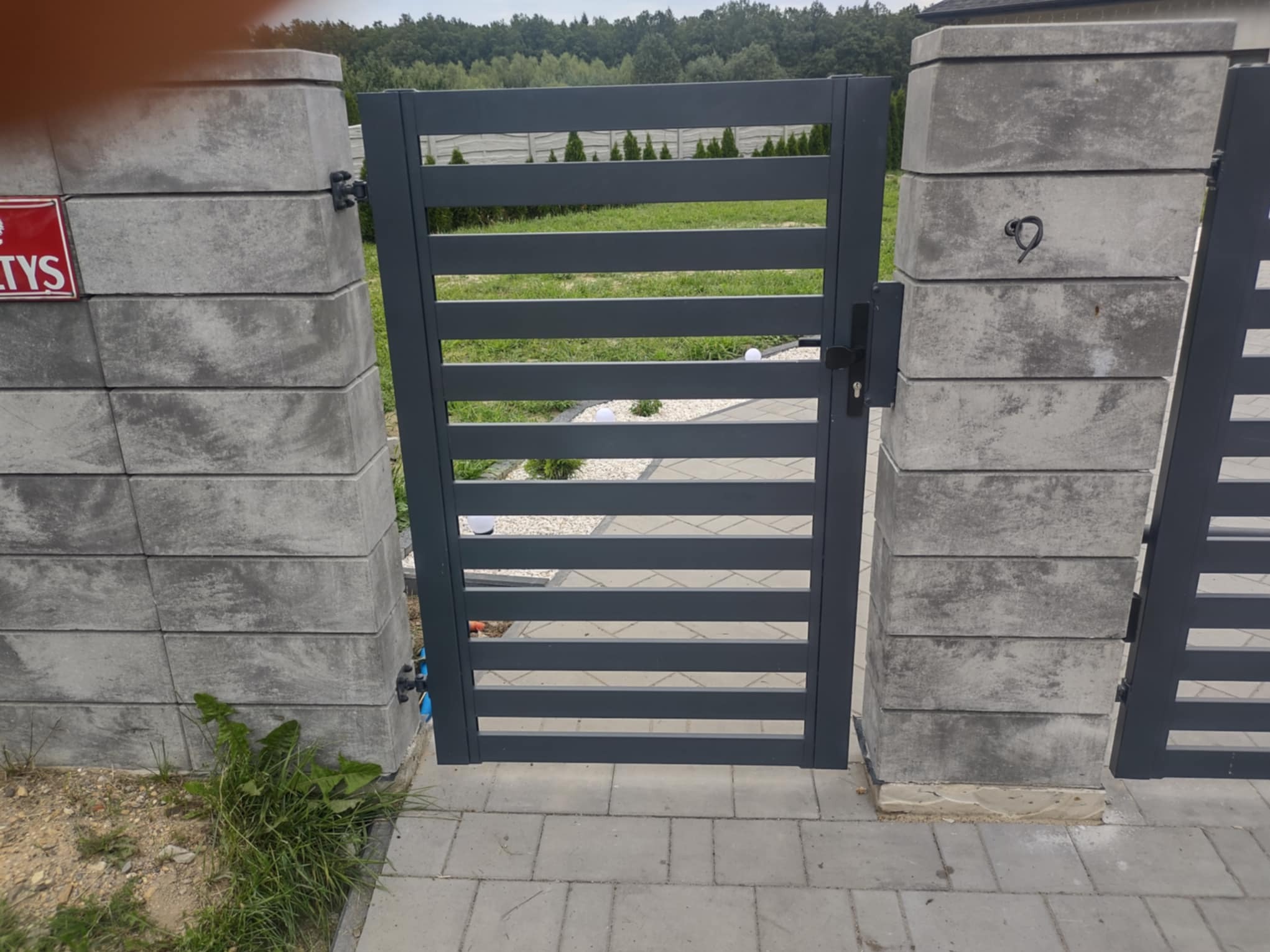 Nowoczesna, szara furtka z poziomymi szczeblami, osadzona na betonowych słupkach z elementami wykończeniowymi, widok z zewnątrz na posesję.