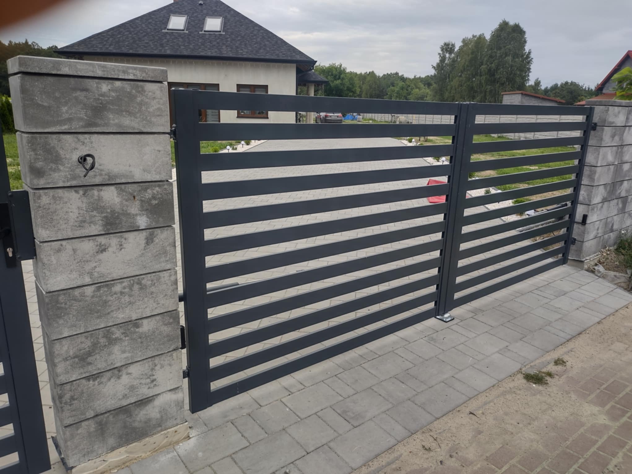 Nowoczesna, grafitowa brama posesyjna z poziomymi panelami, osadzona na betonowych słupkach z szarej kostki, na tle domu z ciemnym dachem i szarej kostki brukowej.