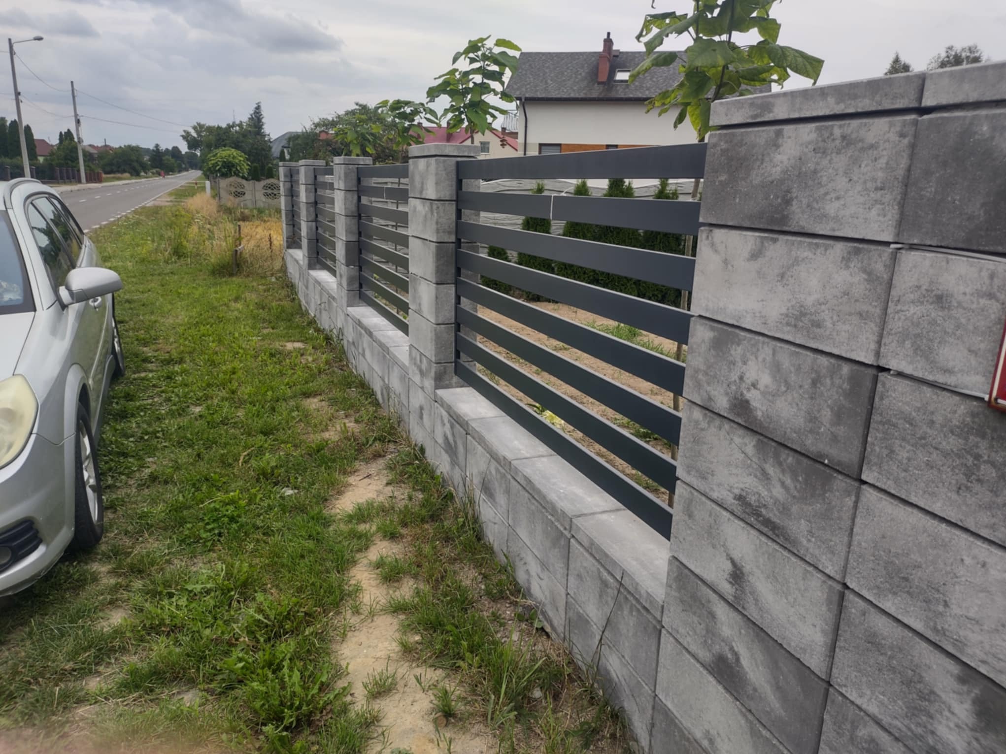 Nowoczesne ogrodzenie z szarych, betonowych bloczków i poziomych, metalowych przęseł, oddzielające posesję od ulicy, z widocznym fragmentem zaparkowanego auta.