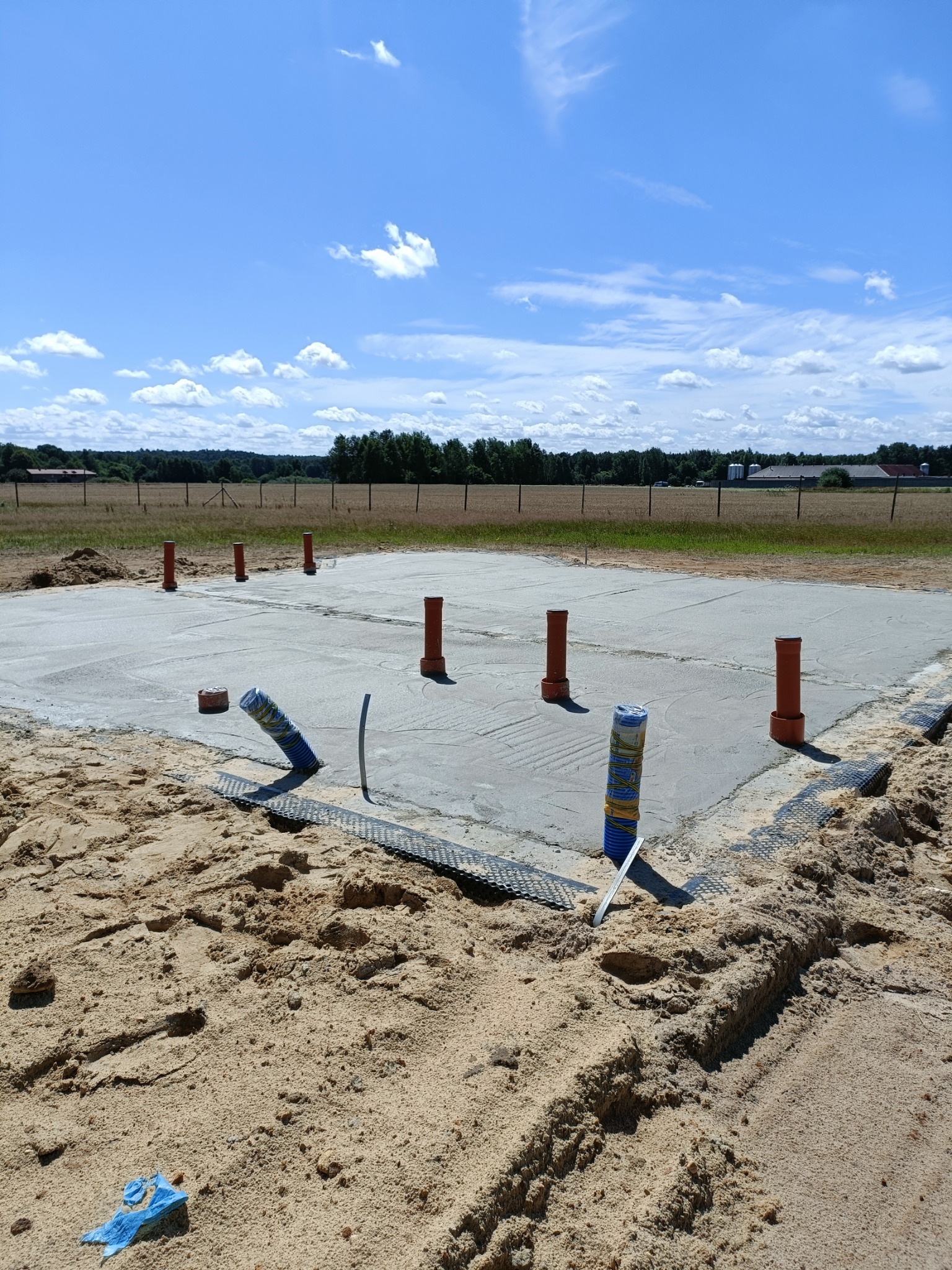 Zalany fundament z wystającymi rurami wentylacyjnymi, otoczony piaskiem i zabezpieczony membraną. W tle łąka i błękitne niebo.