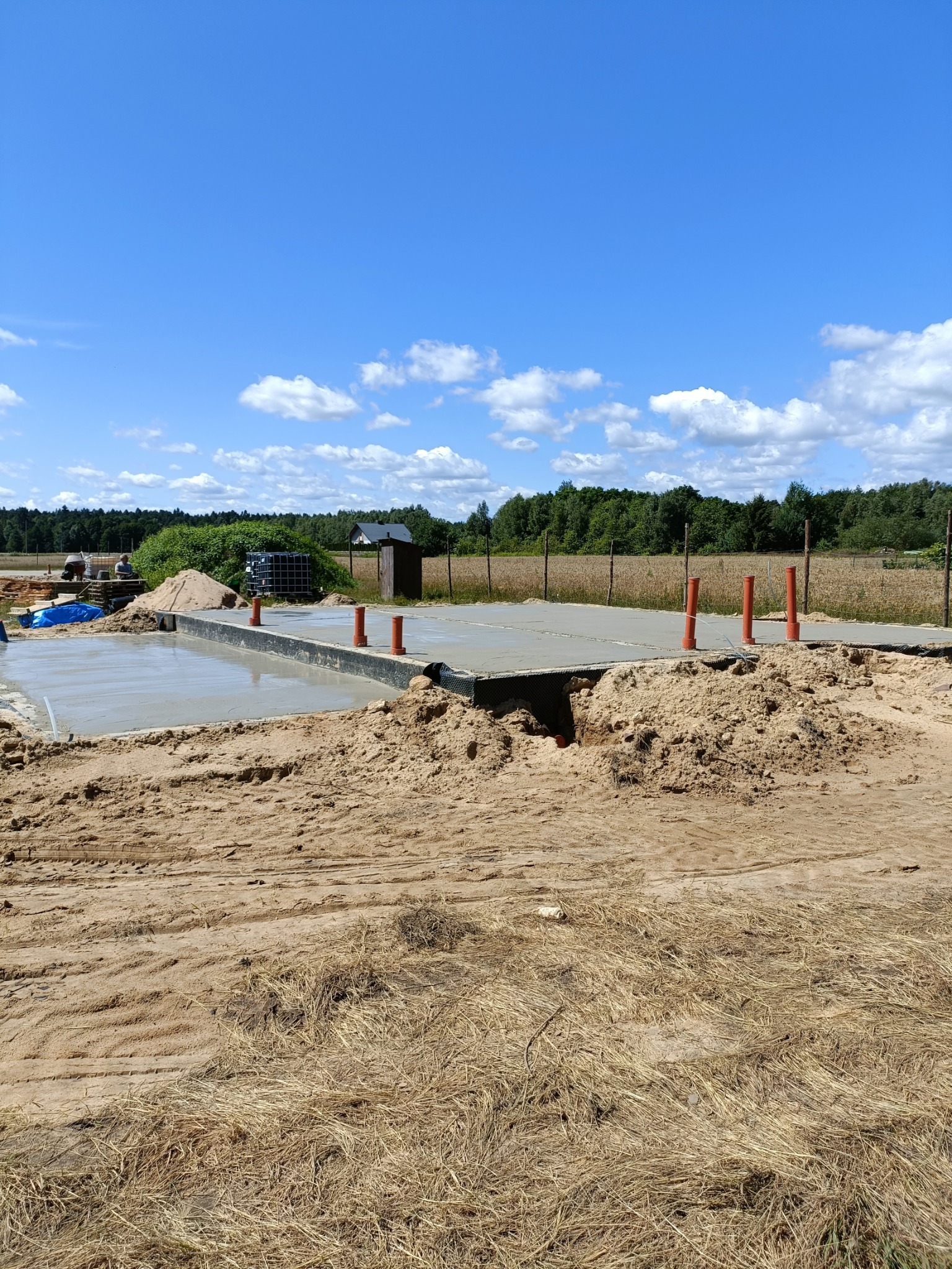 Wylany fundament pod dom z wystającymi rurami wentylacyjnymi na tle błękitnego nieba z chmurami. Widoczny piasek i pracownicy w oddali.